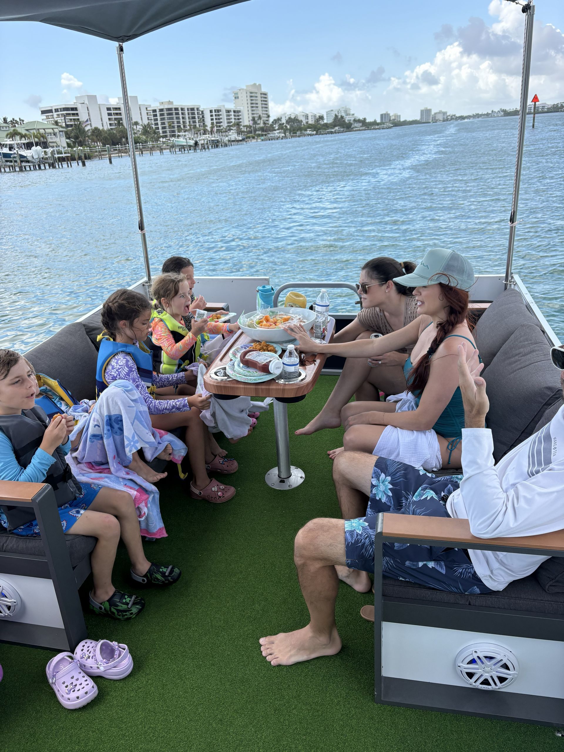 People relaxing on a boat around a table with drinks and snacks, overlooking calm blue water and a city skyline.