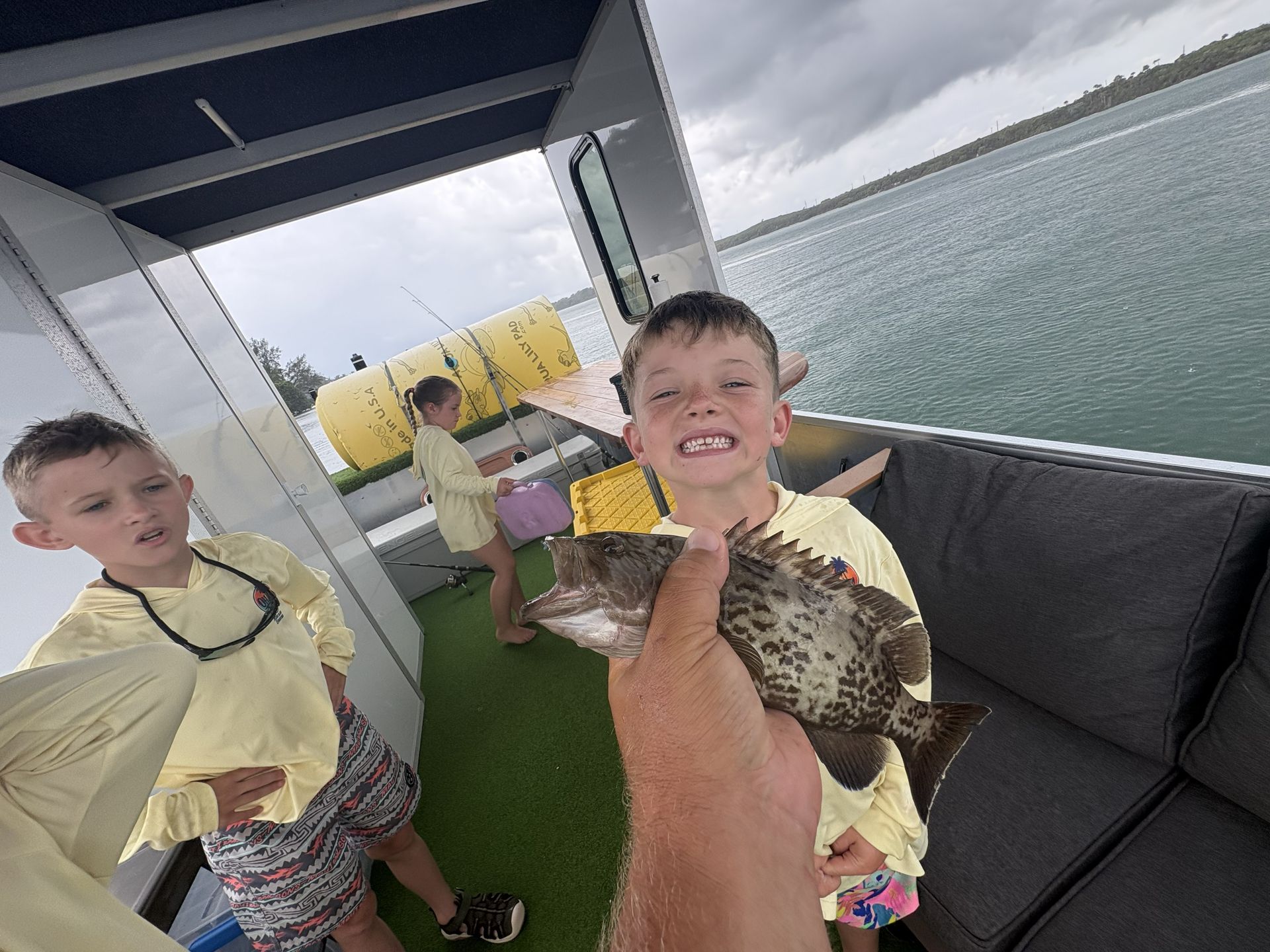 Two boys on a boat deck, one holding a fish, with water and a dock in the background.