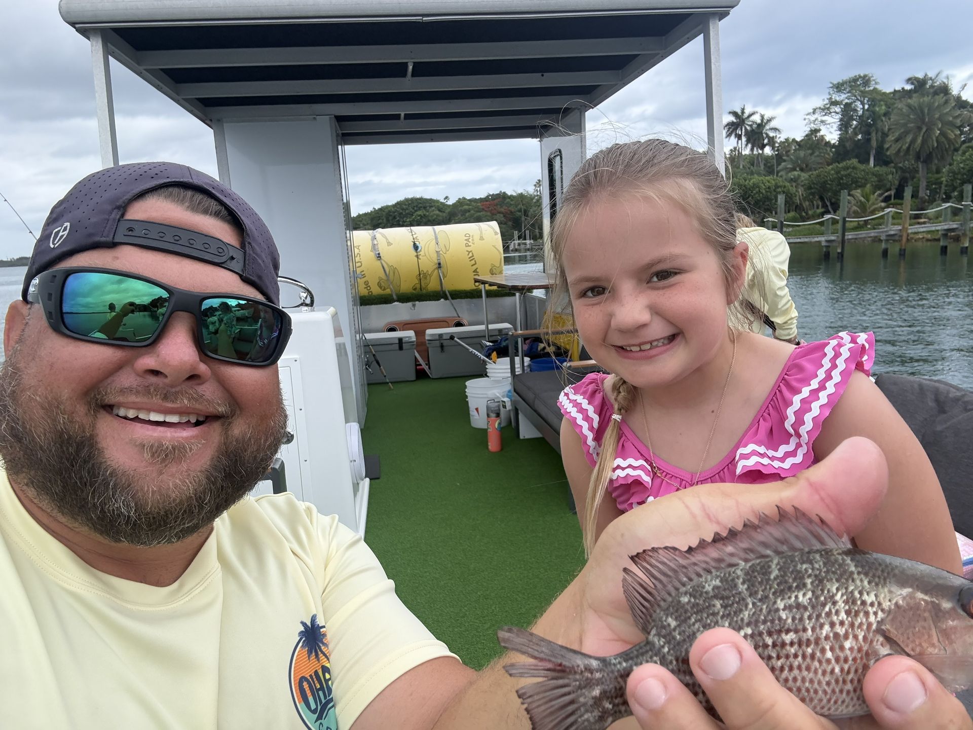 Man and smiling girl on a boat deck holding a fish, with green flooring and water tanks behind them