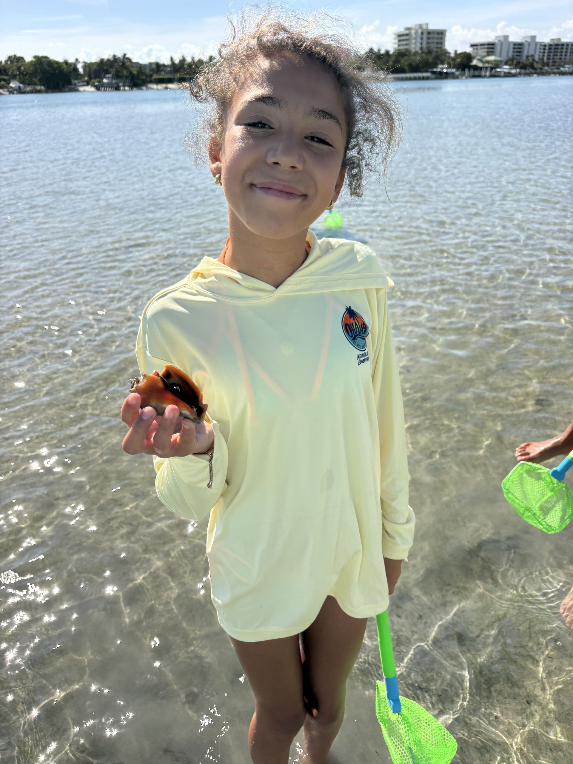 Smiling child standing in shallow water by a lake, holding a shell and a green sand scoop.