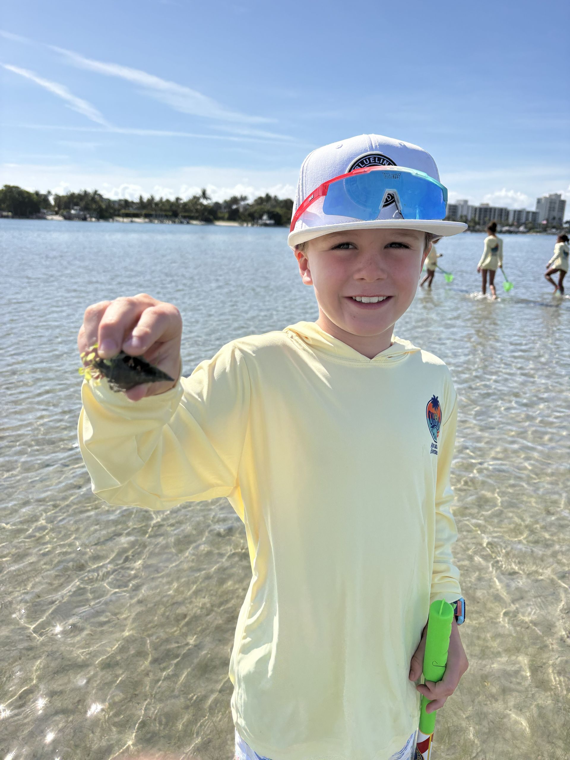 Child standing in shallow water at a beach, smiling and holding up a small shell in a yellow shirt and white cap