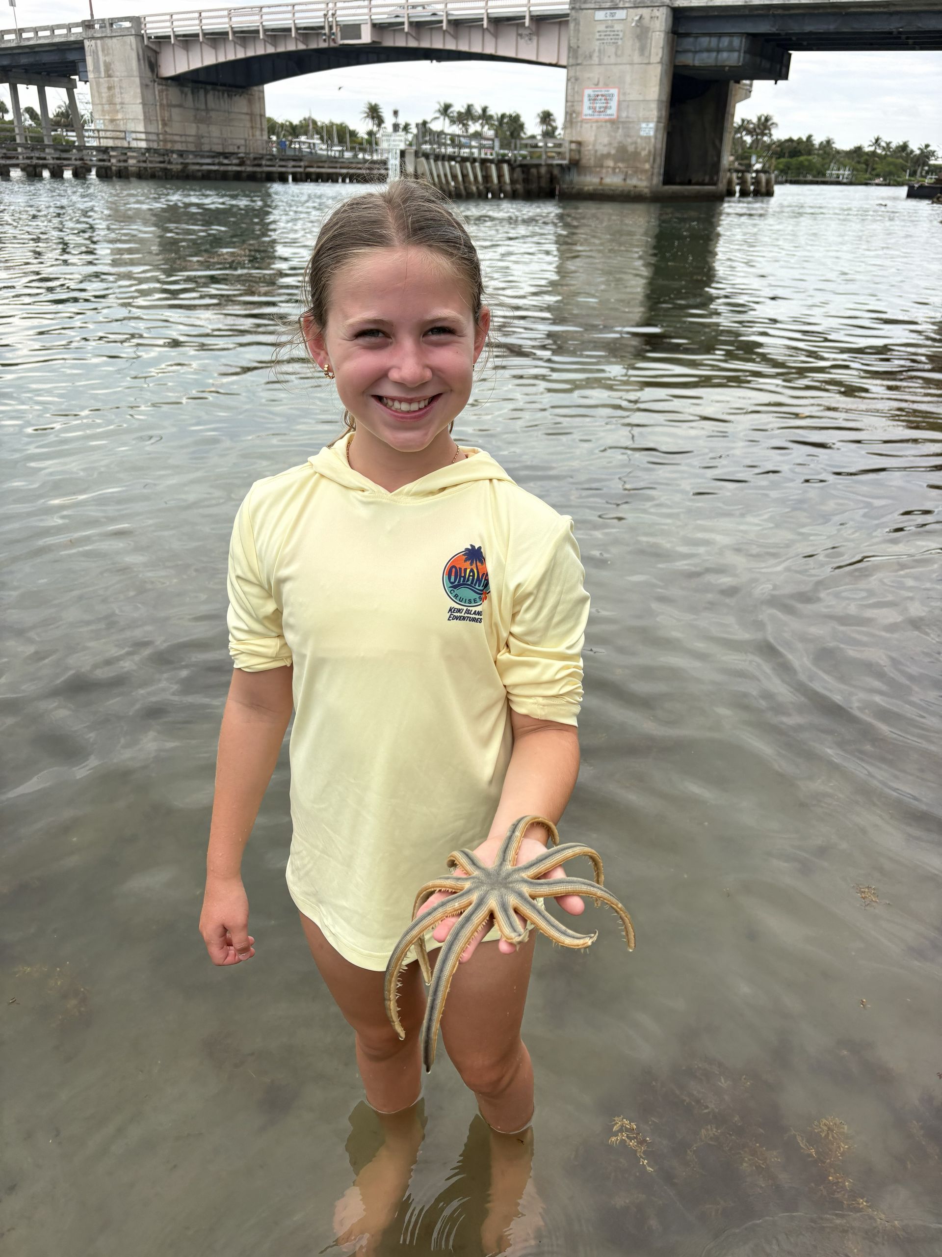 Smiling child in a pale yellow shirt holding a starfish while standing in shallow water near a bridge