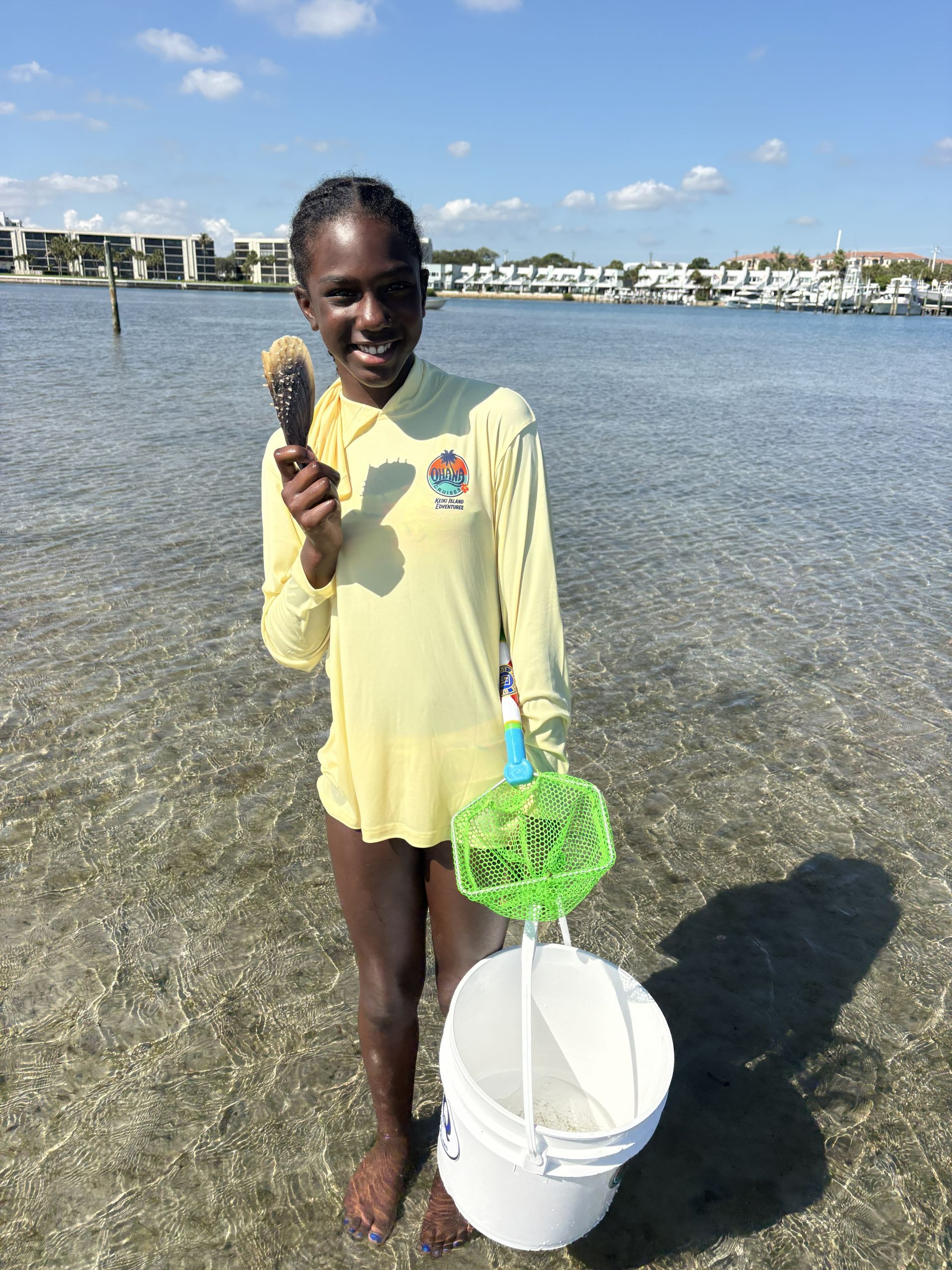 Person standing in shallow water holding a scoop and bucket with green netting, smiling near the shore