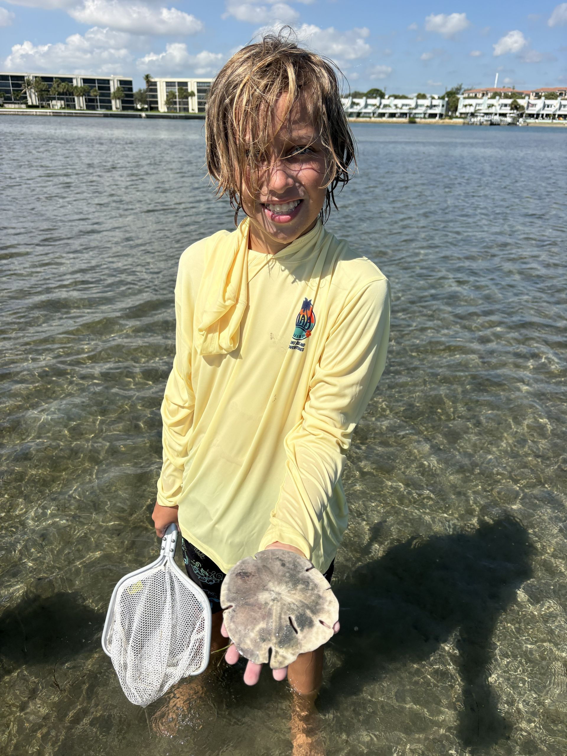 Child standing in shallow water holding a white net and a seashell, with waterfront buildings in the background