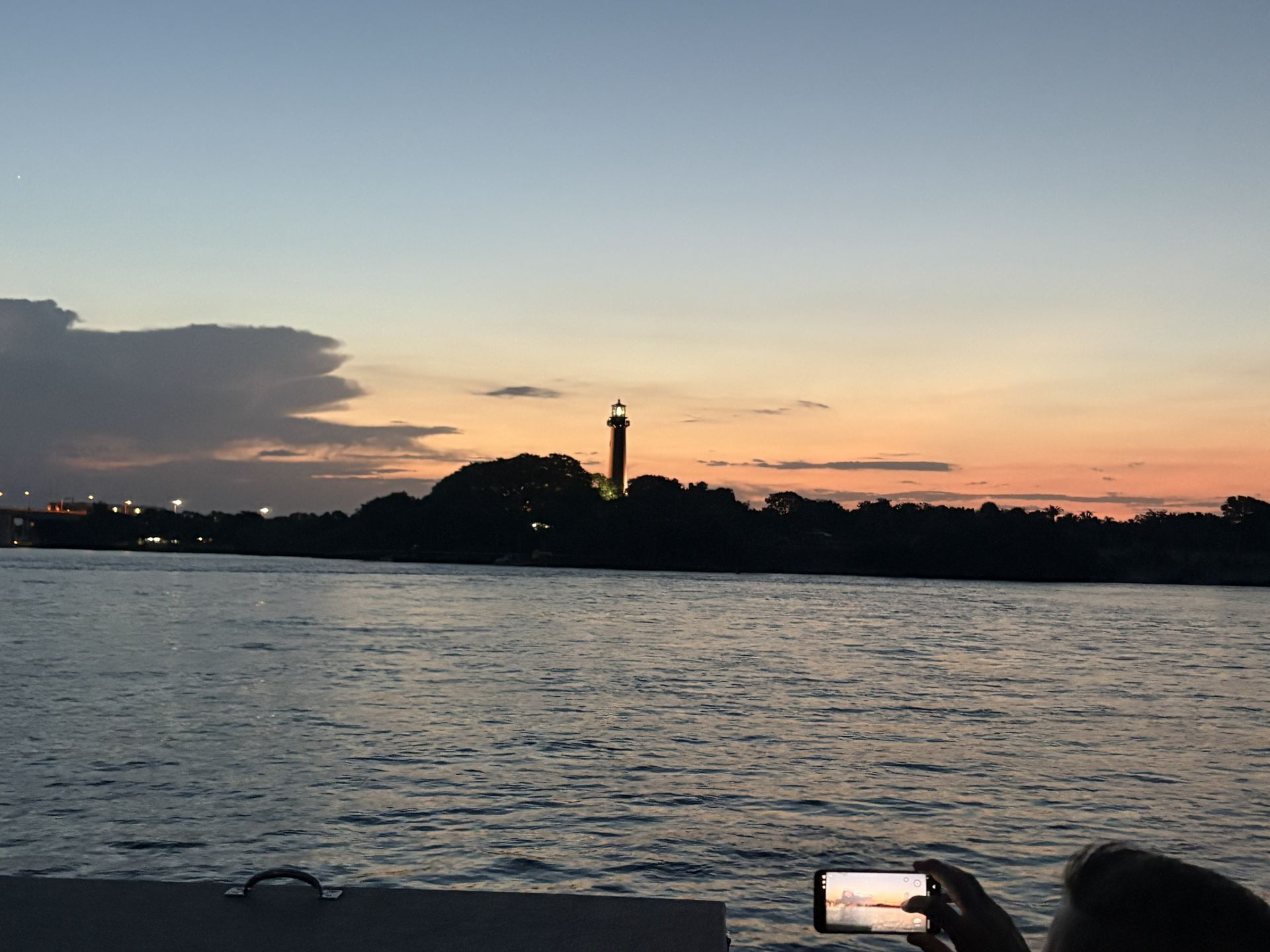 Sunset over water with silhouetted shoreline and lighthouse; phone screen glowing in the foreground