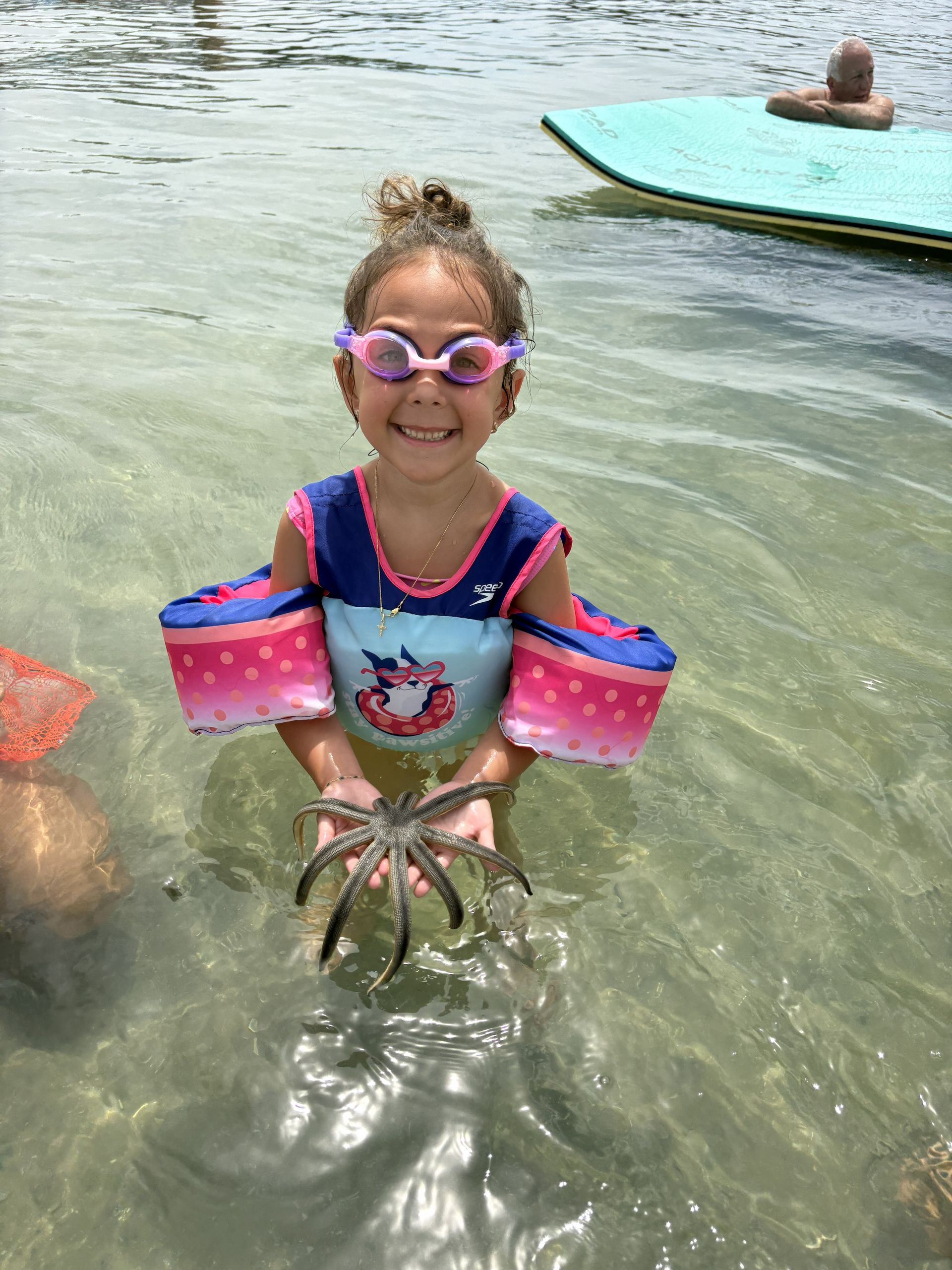 Child in purple goggles and pink arm floaties stands in shallow water, smiling near a floating mat.