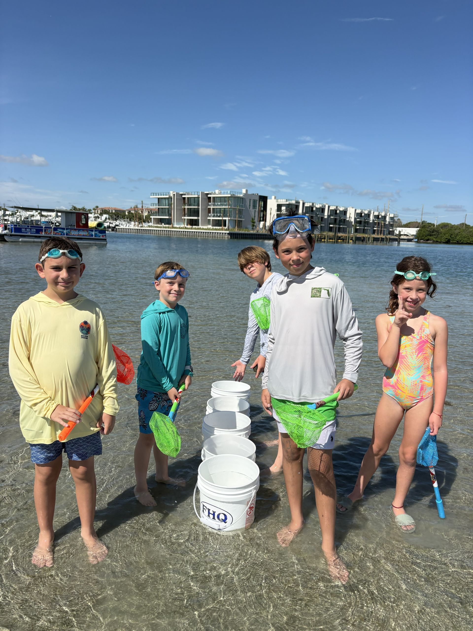 Five children in swimwear stand in shallow water holding buckets on a sunny beach.