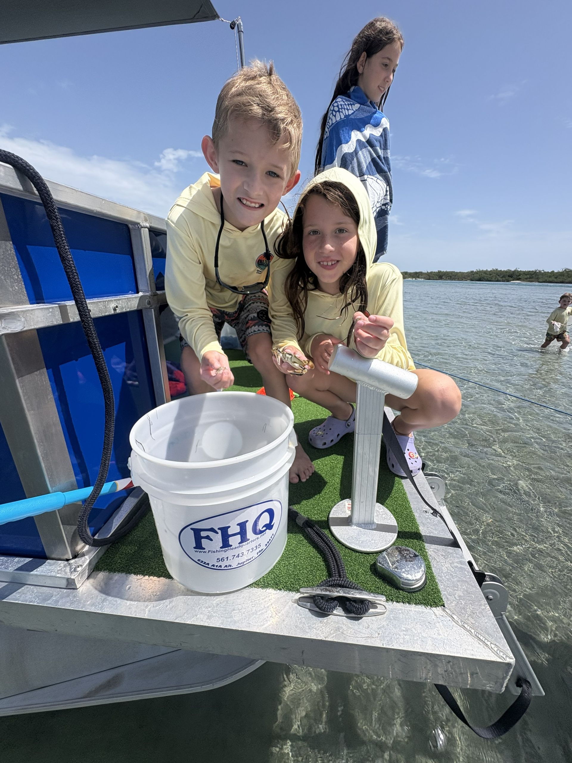 Two children smiling on a boat deck, holding a fishing net and a white bucket on the water.