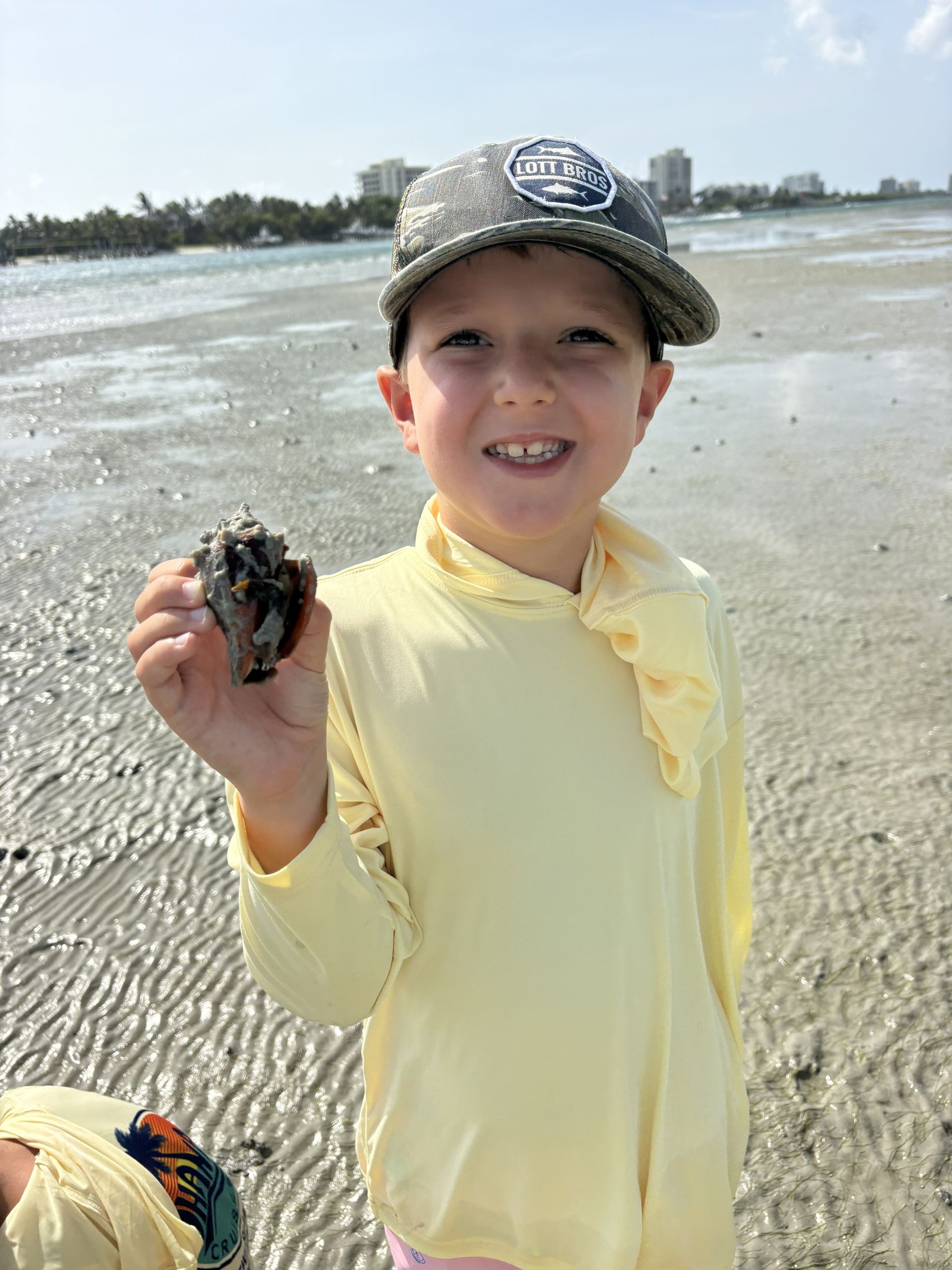 Child on a beach holding a small crab, wearing a yellow shirt and cap.