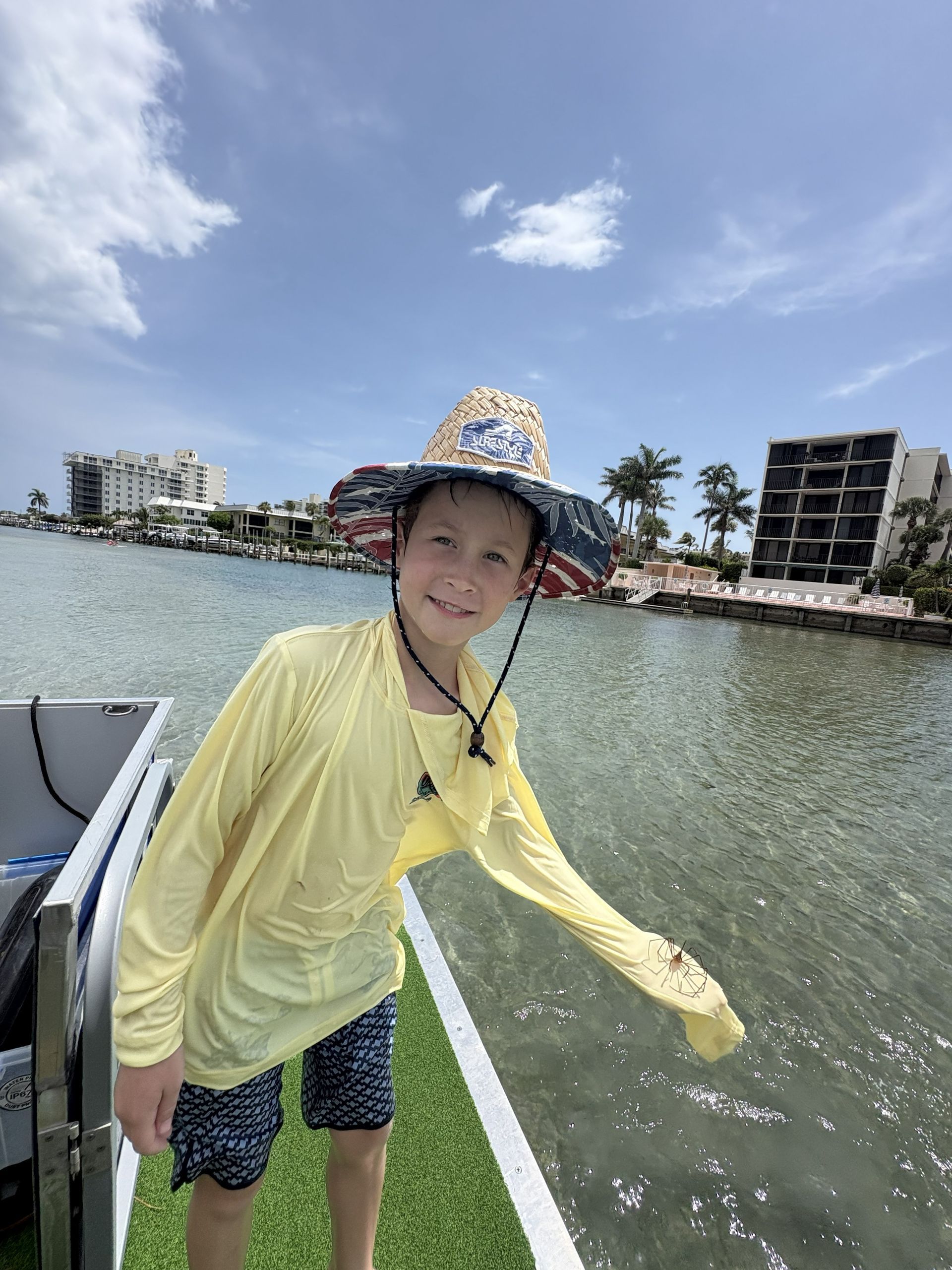 Person in yellow rain gear and patterned hat smiling on a dock by the water under a blue sky.