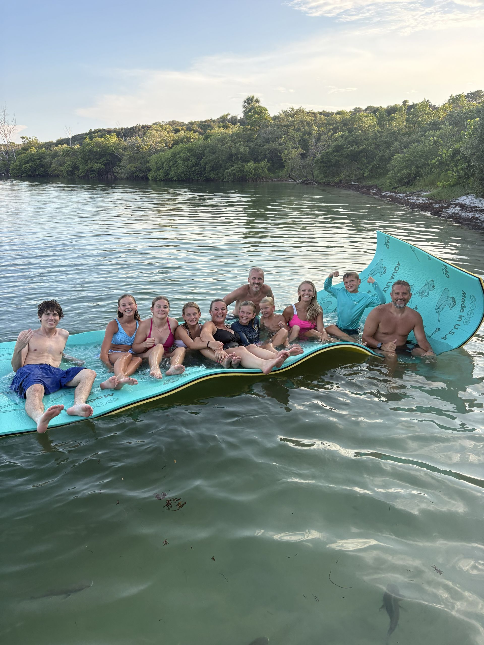 Group lounging on a floating raft in a lake beside a wooded shoreline at sunset