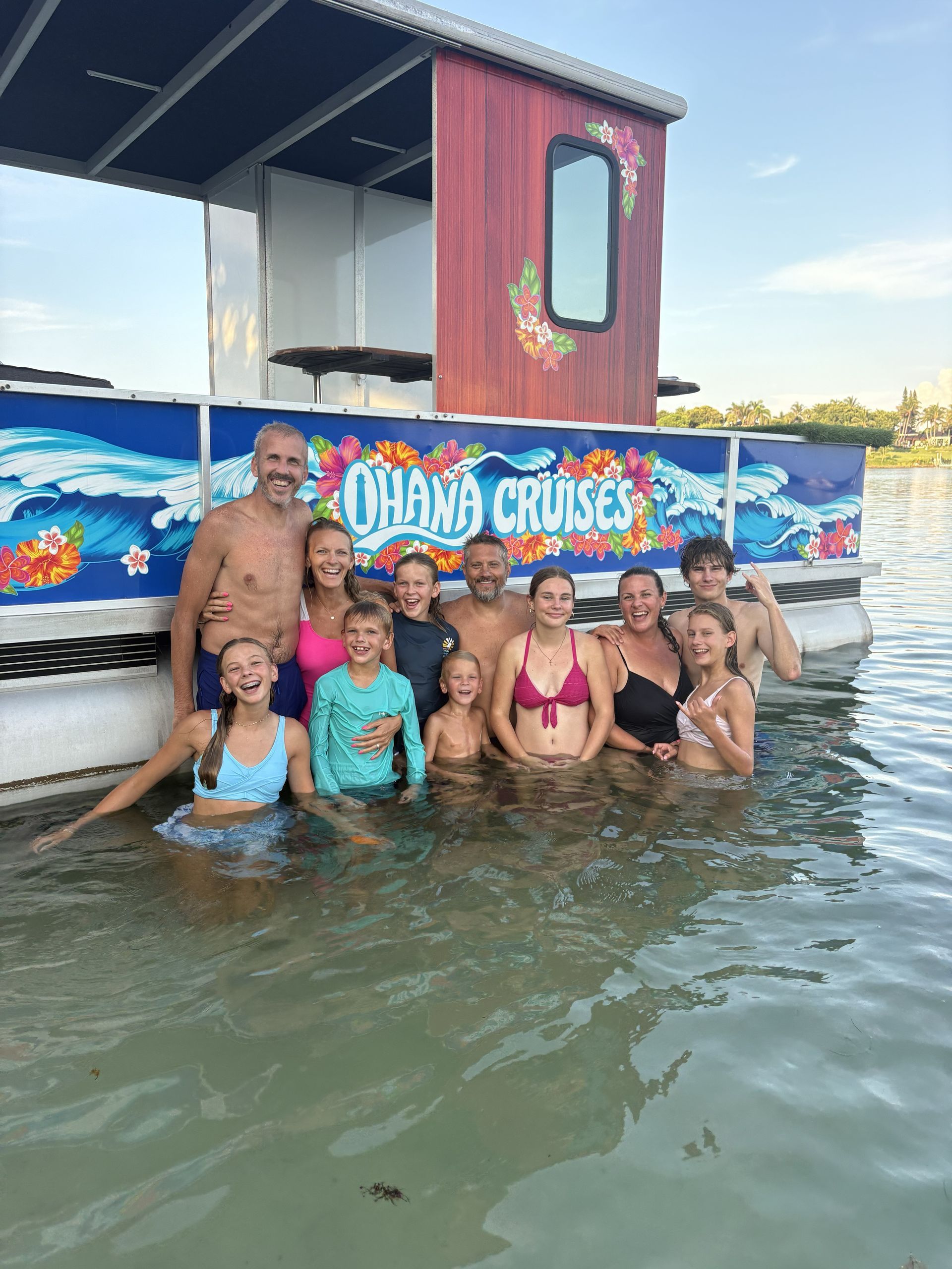 People posing in shallow water beside a colorful “Chama Cruises” boat at sunset