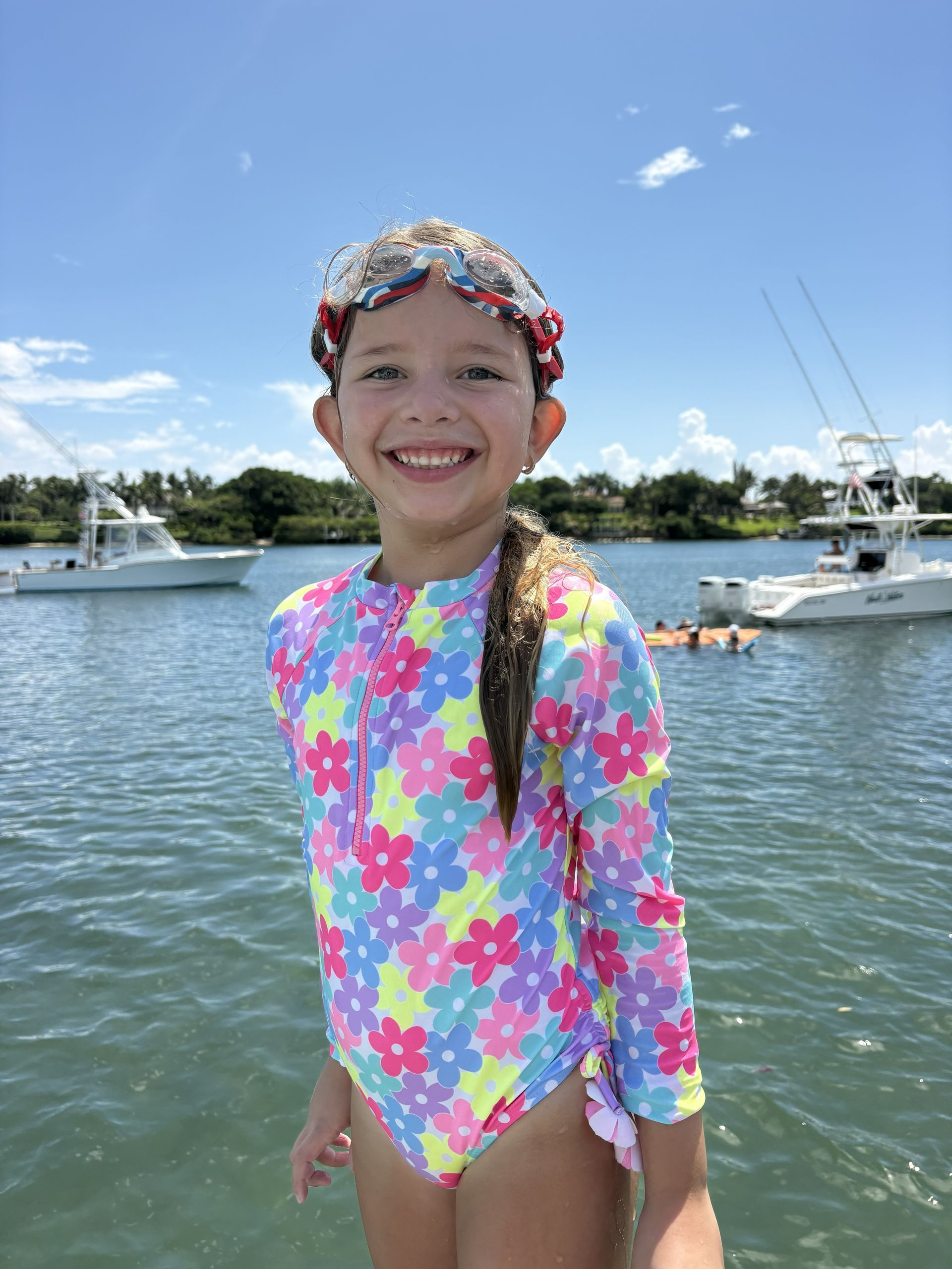 Child in a bright floral swimsuit smiles at a marina with boats and calm water under a blue sky.