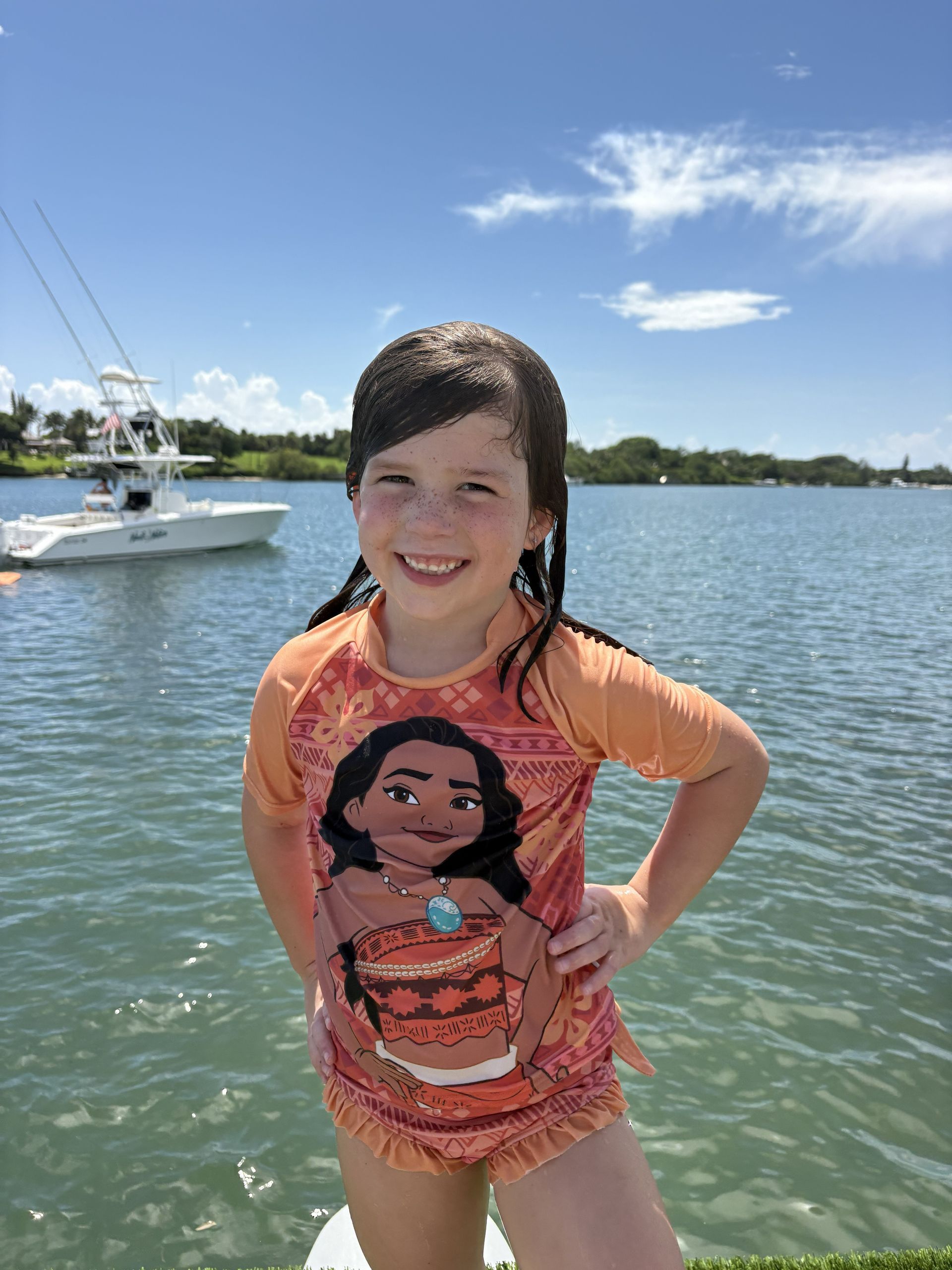 Smiling child in an orange swimsuit standing in shallow water near a boat on a sunny day