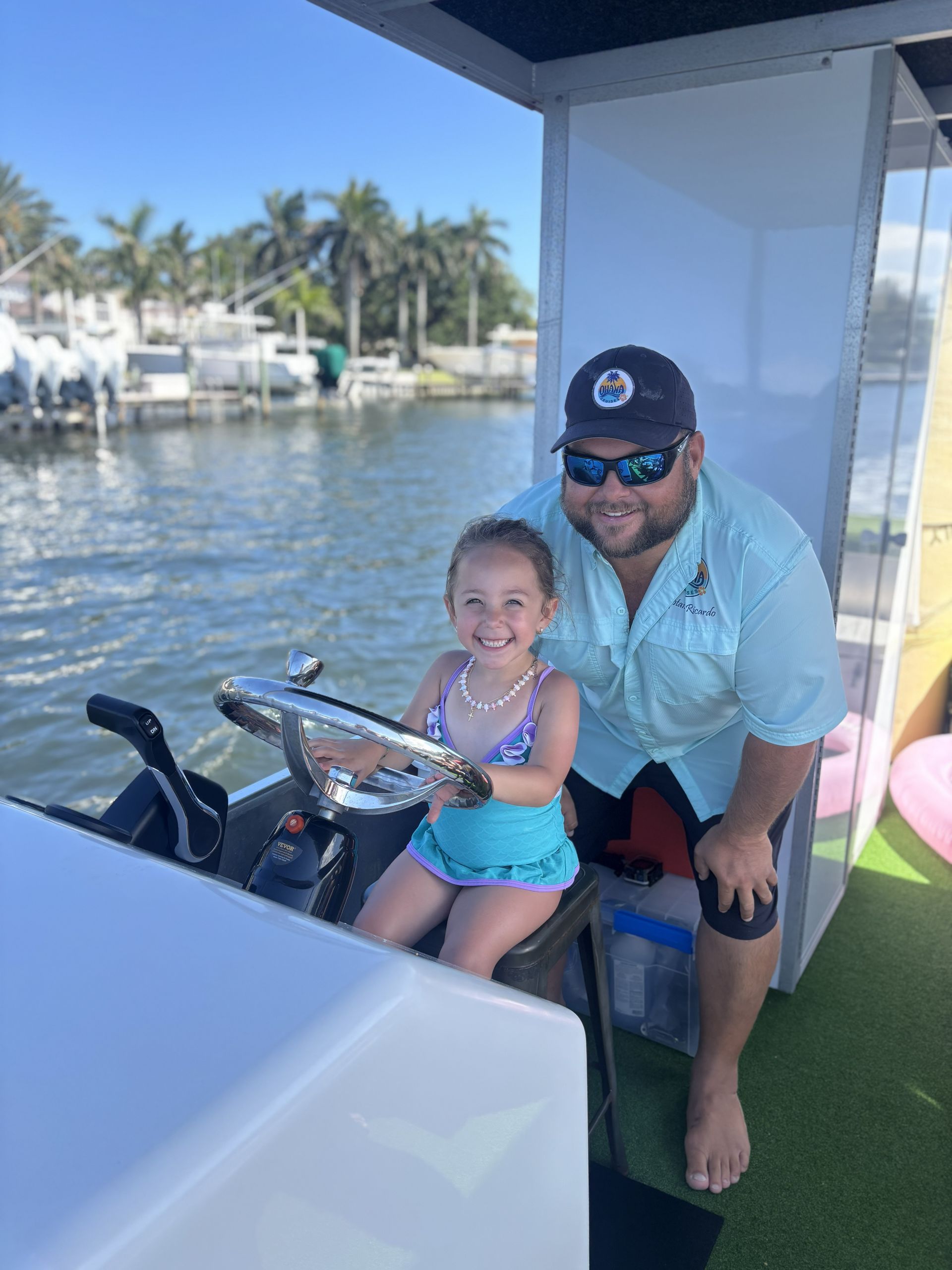 Man and child smiling on a boat near a marina, with calm water and palm trees in the background