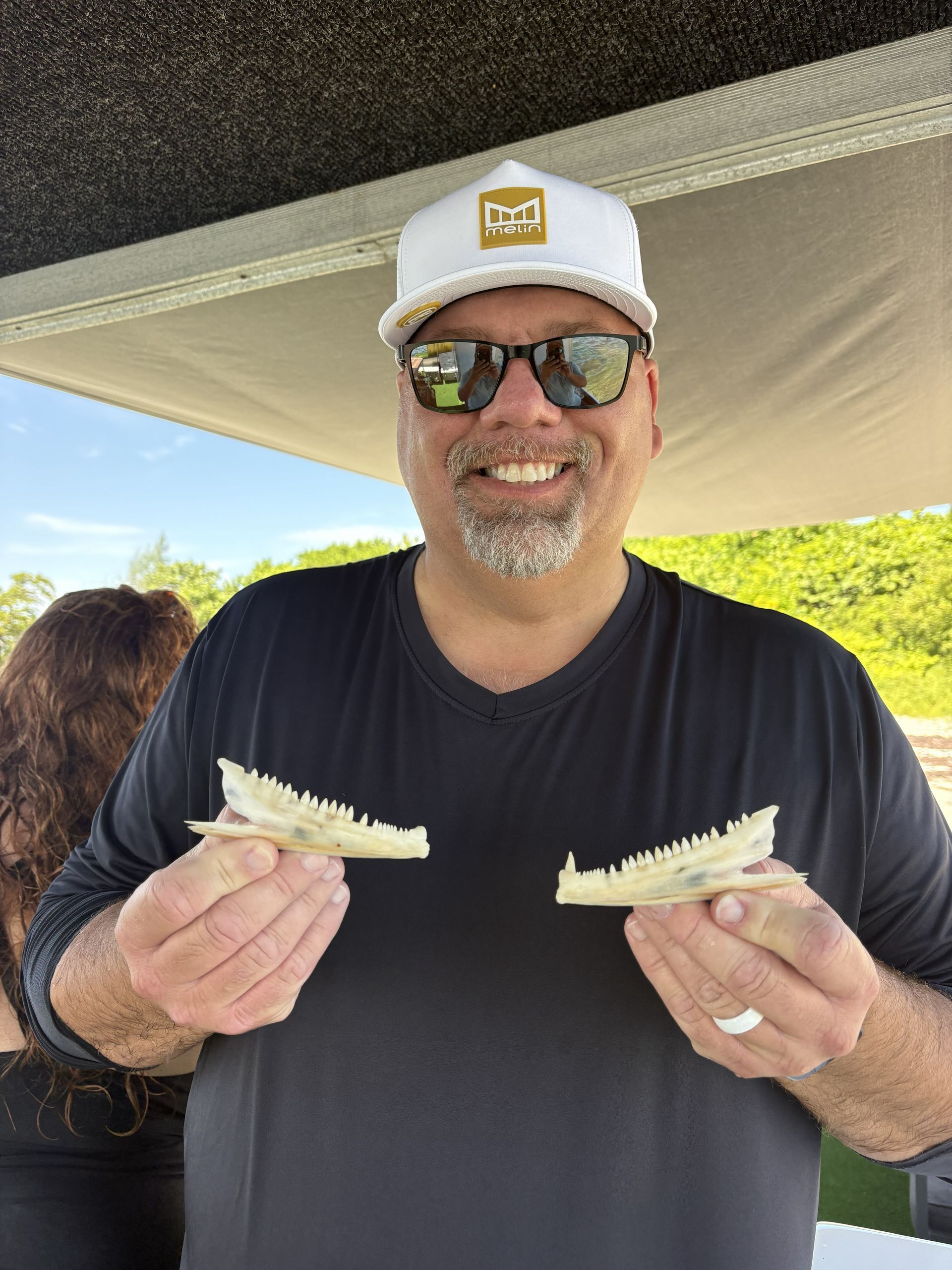 Man smiling outdoors under a canopy, holding two sandwich halves and wearing a white cap and sunglasses