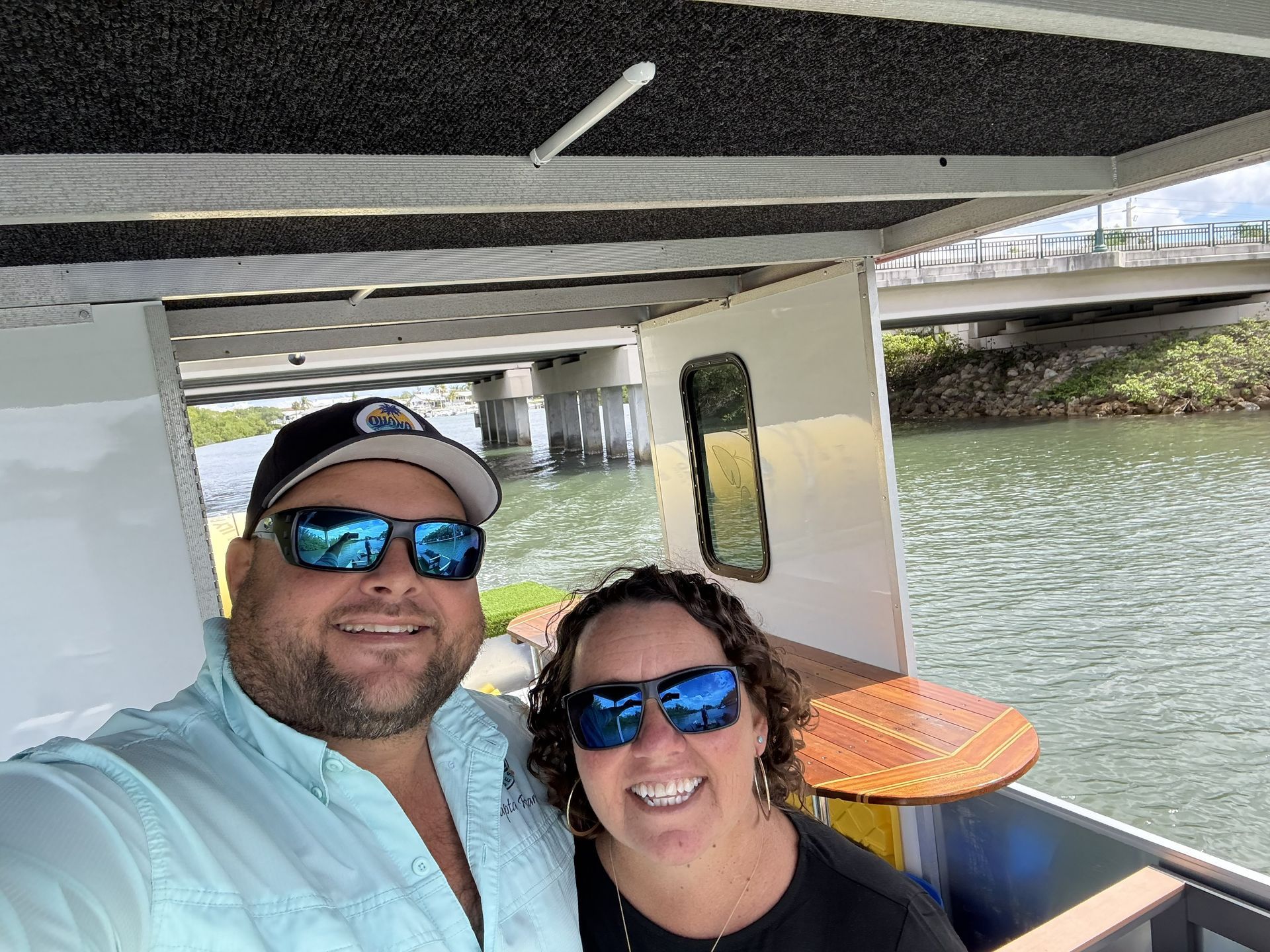 Two smiling people wearing sunglasses on a covered boat ride with water in the background