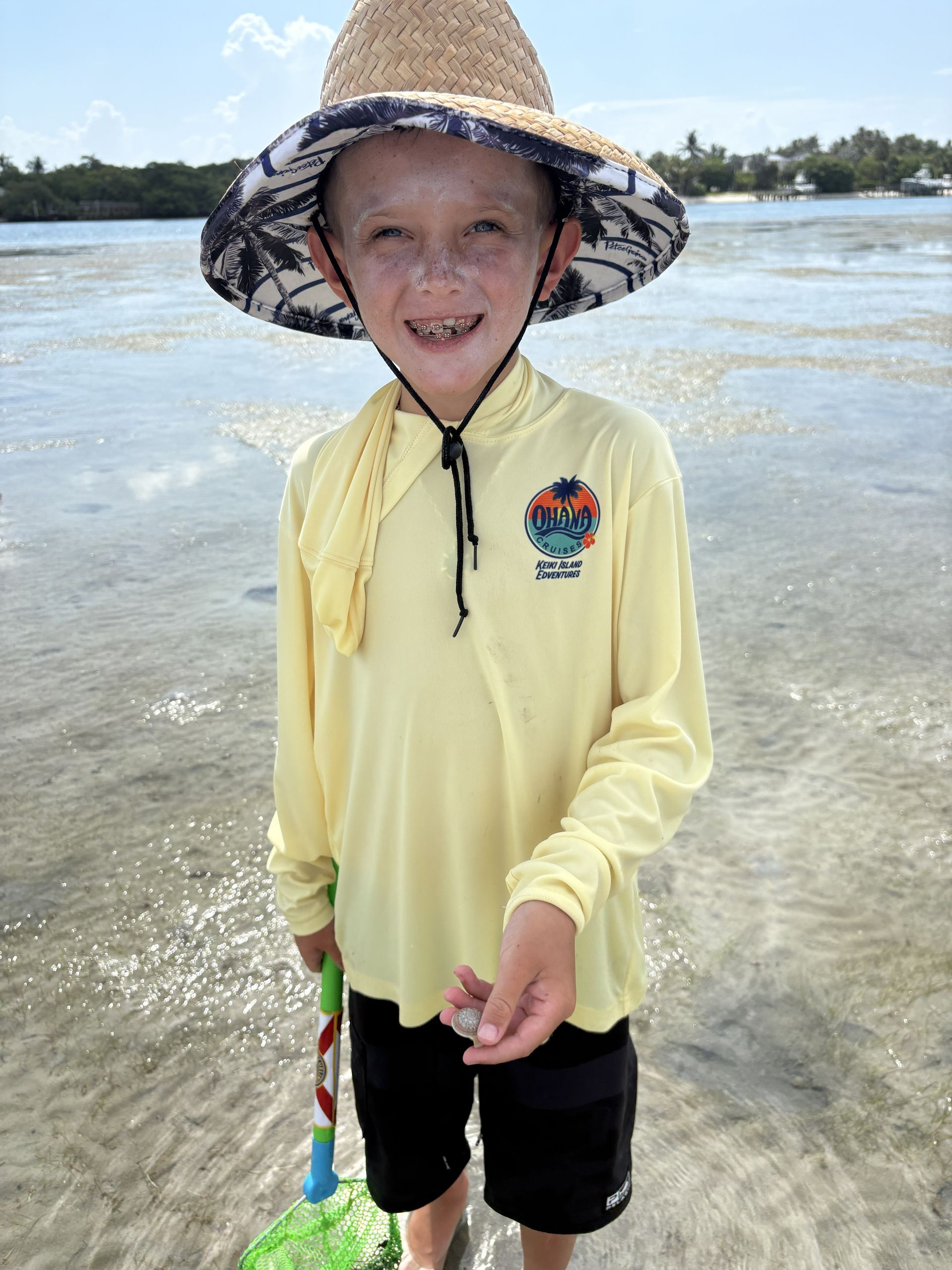 Child standing in shallow water wearing a straw hat, yellow shirt, and black shorts, smiling at the beach