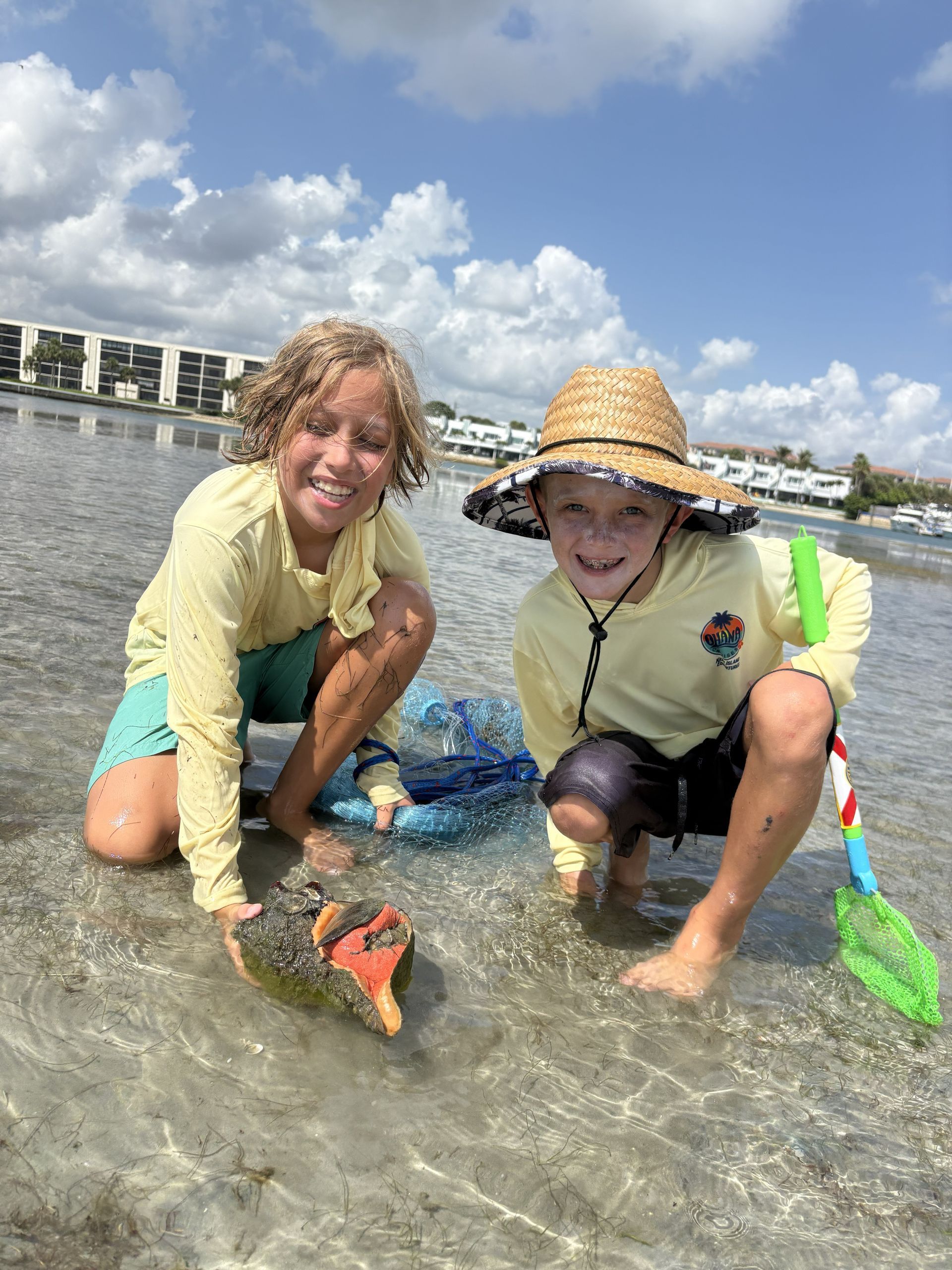 Two children kneel in shallow water on a sunny beach, smiling and holding colorful buckets.
