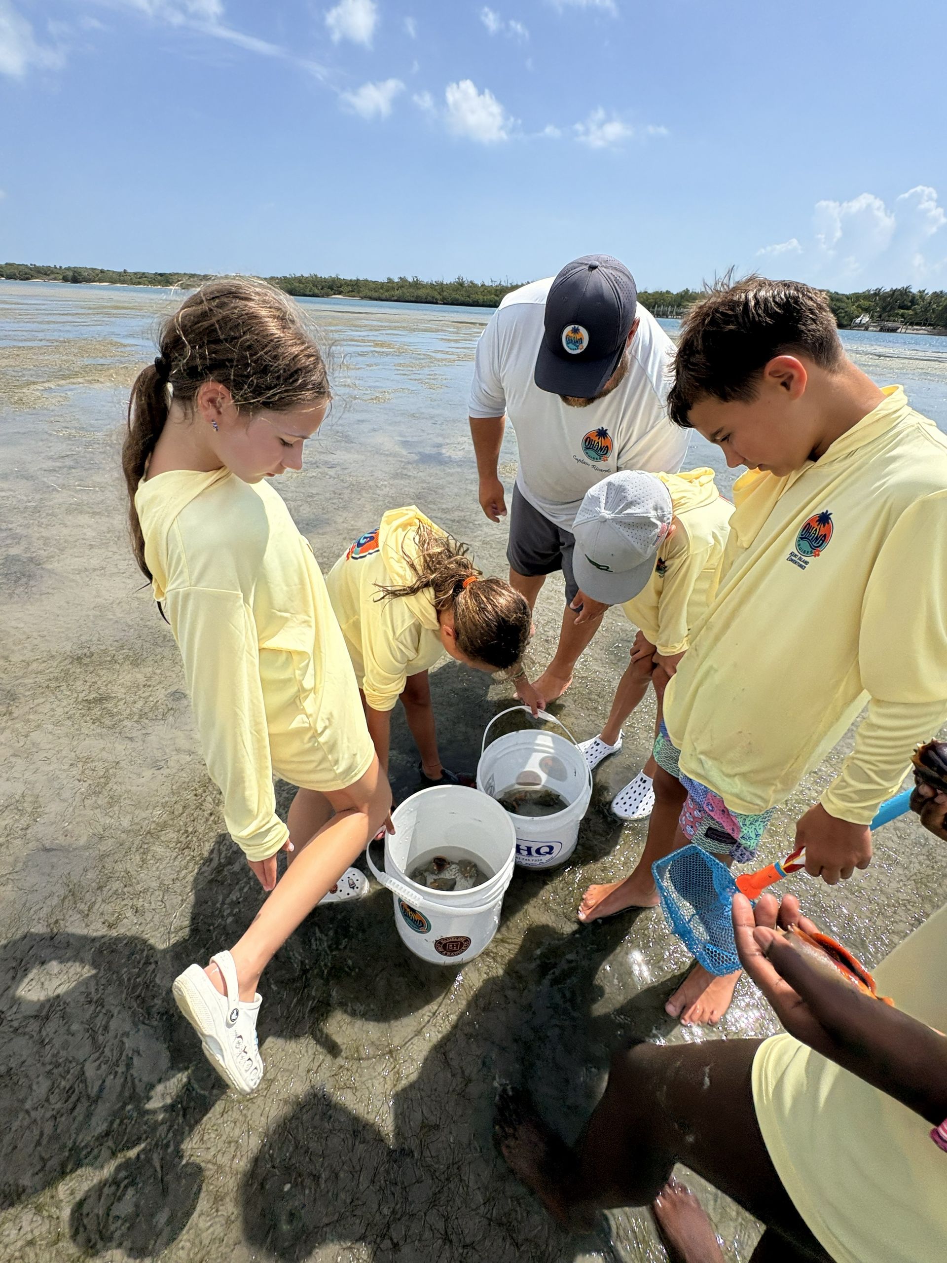 People gathered on a muddy shoreline, using white buckets and nets to inspect collected samples.