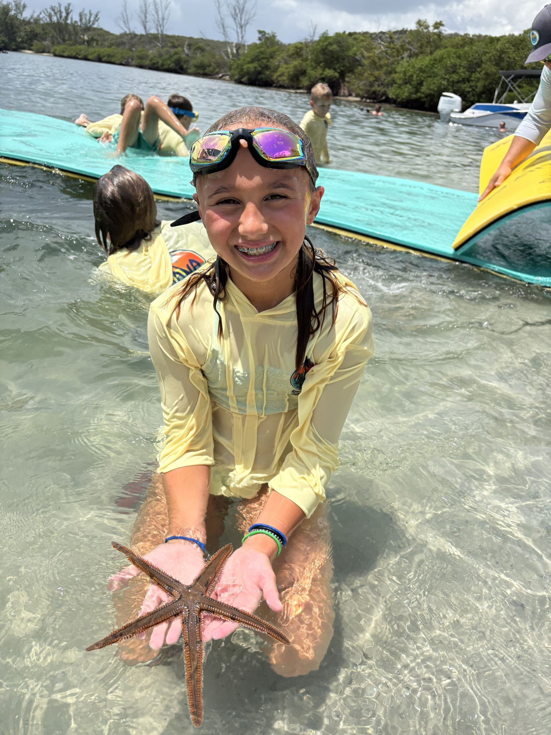 Smiling child in a yellow rash guard holding a starfish in shallow clear water by a sandy beach
