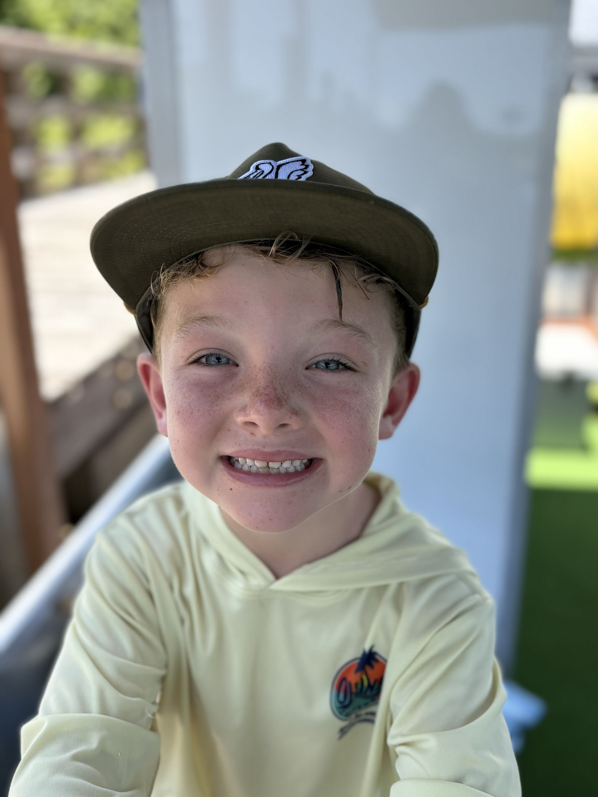 Smiling child in a cap and light hoodie outdoors, standing near a railing.