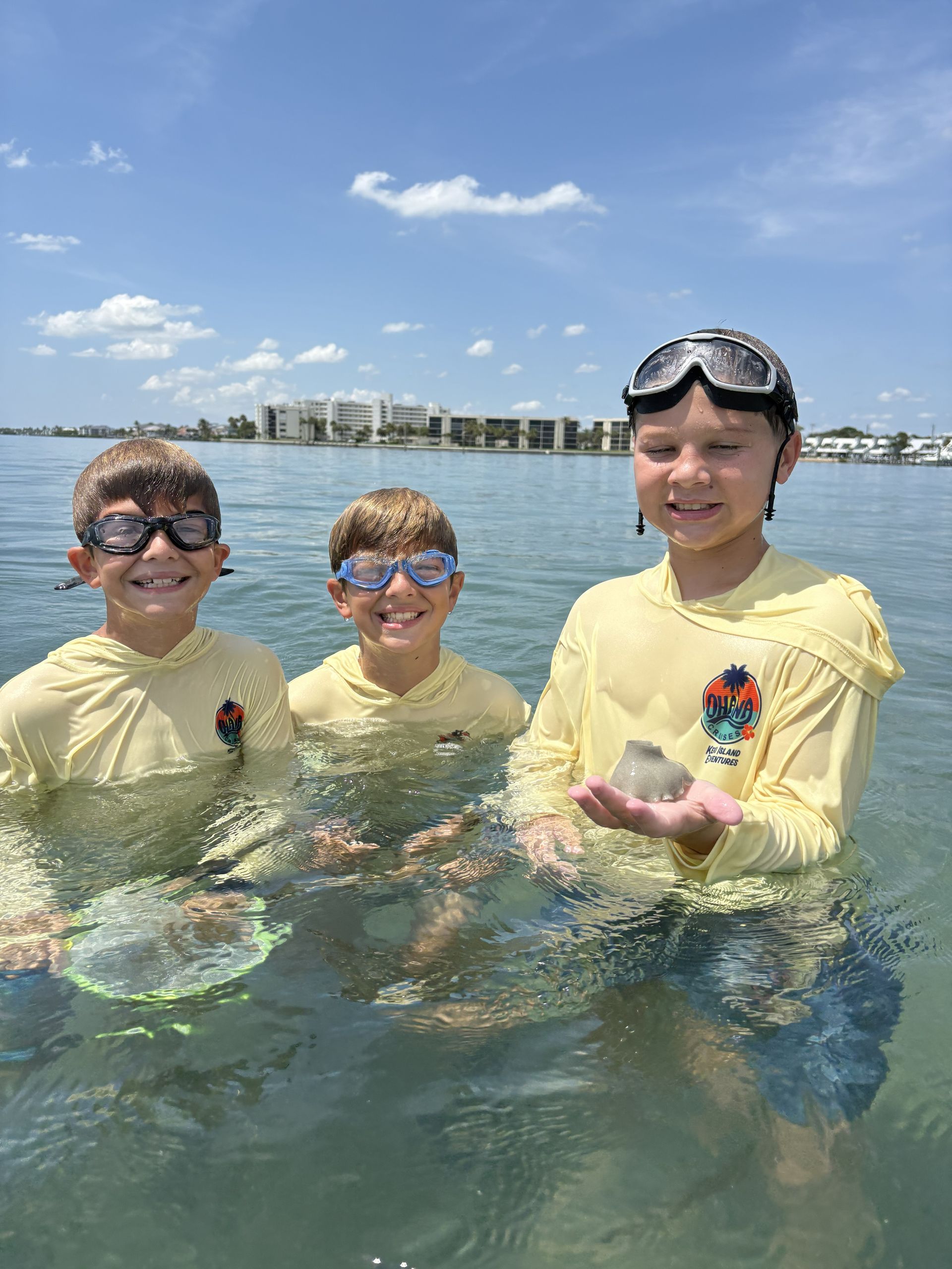 Three people in yellow rash guards standing in shallow water, smiling at the beach under a bright blue sky.