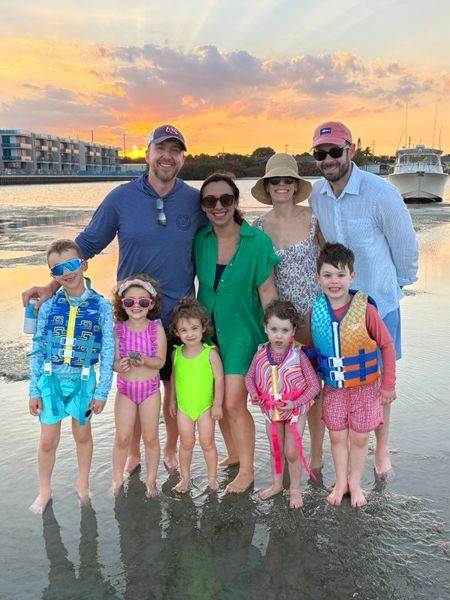 Family standing in shallow beach water at sunset, smiling in colorful swimwear and sunglasses.