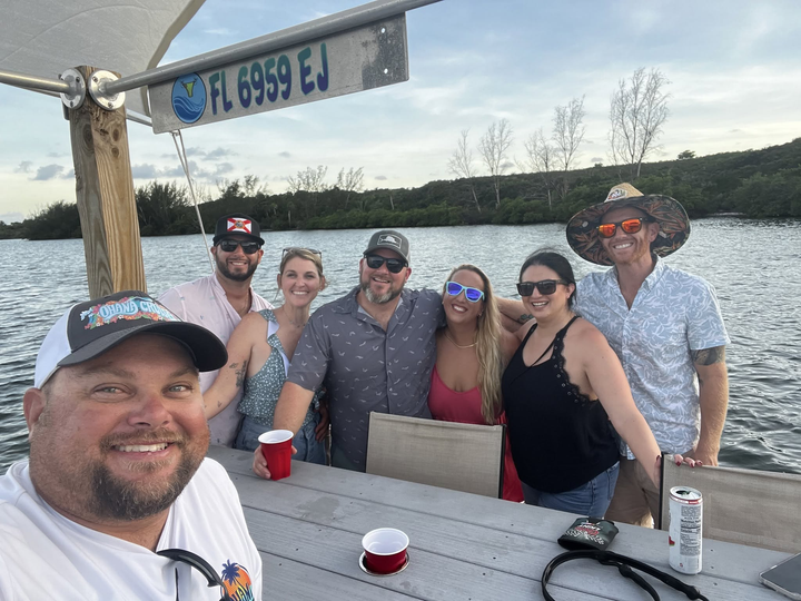 Group of friends posing at a picnic table by a lake, smiling with drinks on a sunny day.