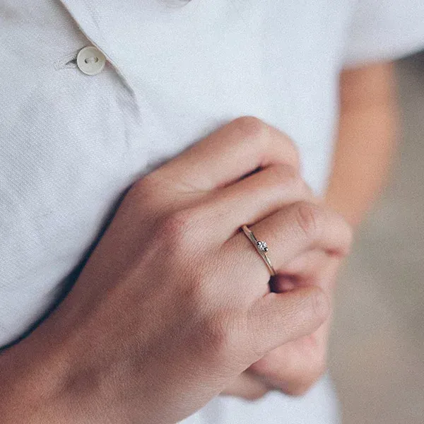 De hand van een persoon die een delicate gouden ring met een klein juweeltje draagt, rustend op een wit overhemd.