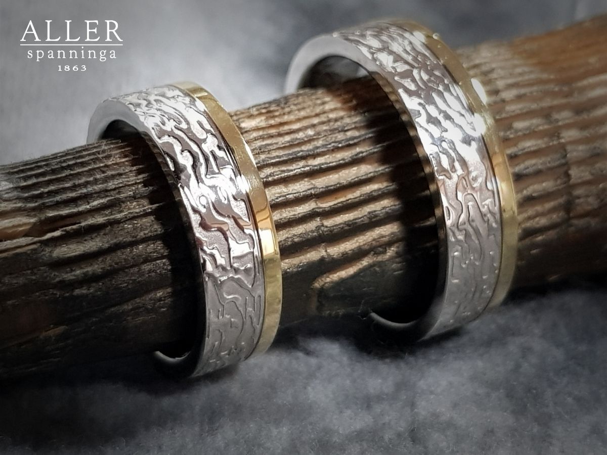 Drie trouwringen van zilver en goud op een houten stokje, met een getextureerd metalen ontwerp.