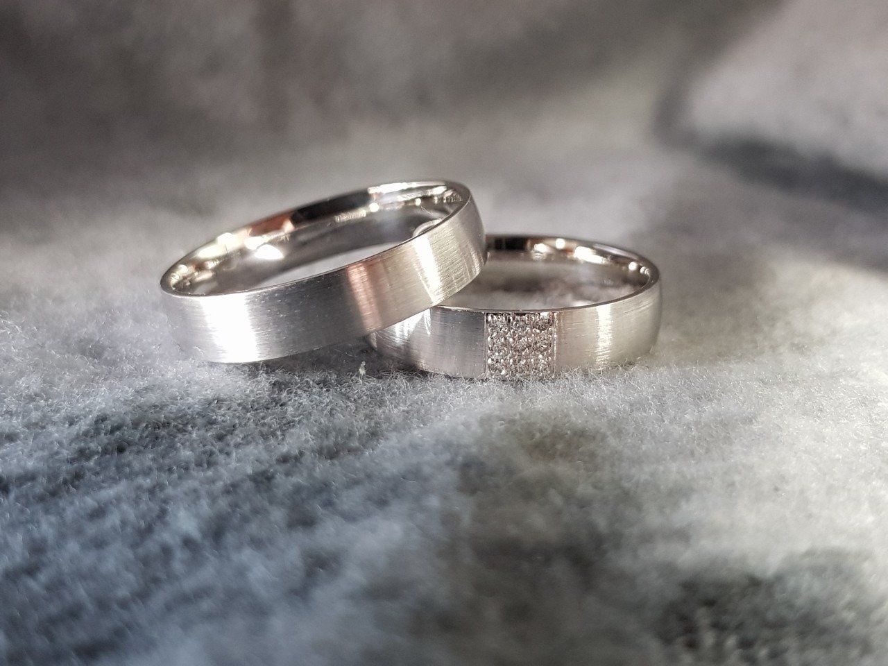 Twee zilveren trouwringen, waarvan één met een klein vierkantje met diamanten, rustend op zachte grijze stof.