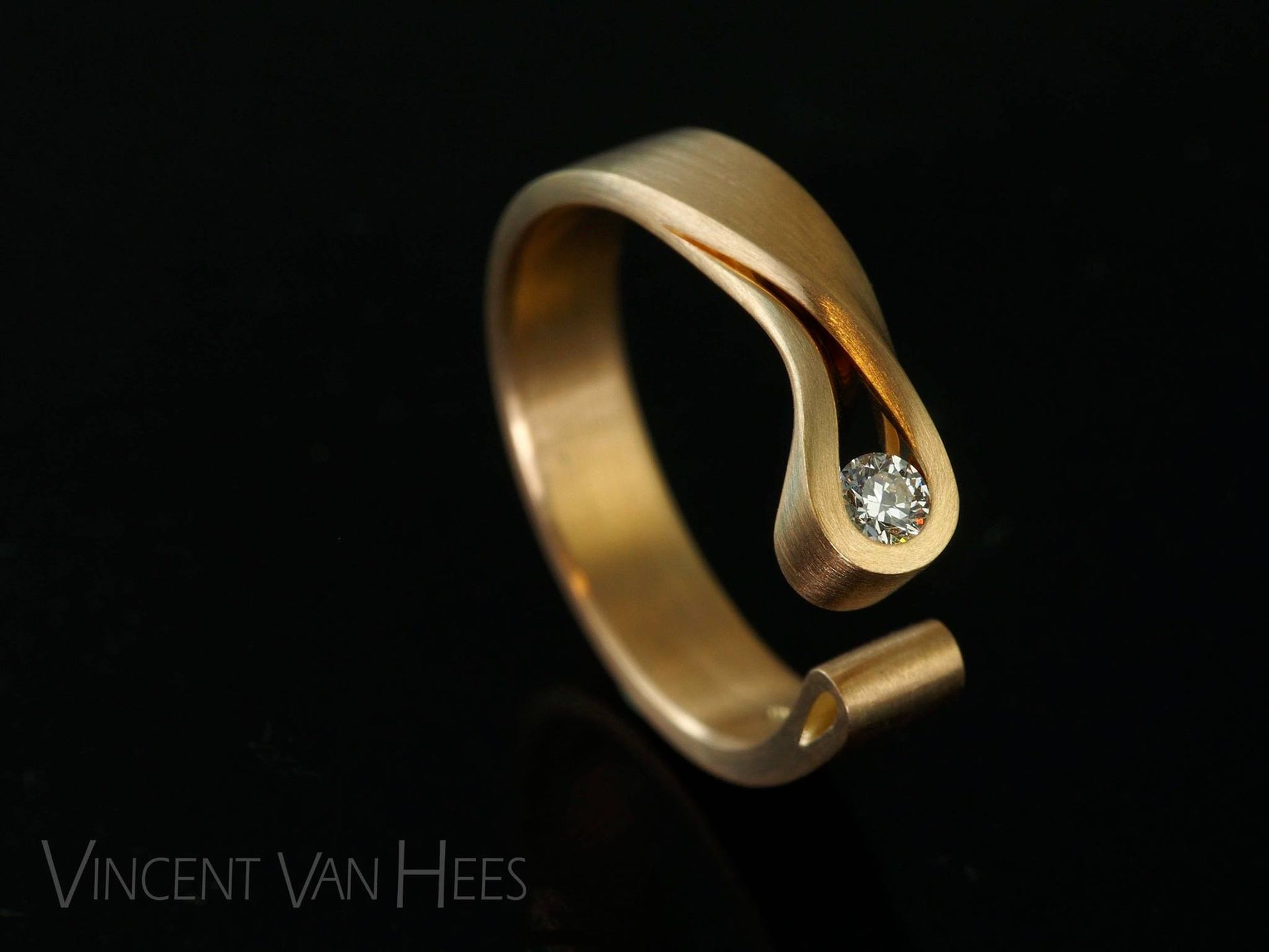 Gouden ring met een diamant, gezet op een zwarte achtergrond. De band loopt gebogen en eindigt in een zetting.