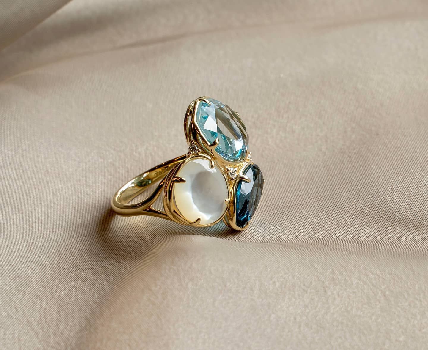 Gouden ring met drie edelstenen: een lichtblauwe ovaal, een witte ovaal en een donkerblauwe ovaal, afgezet op een beige stof.
