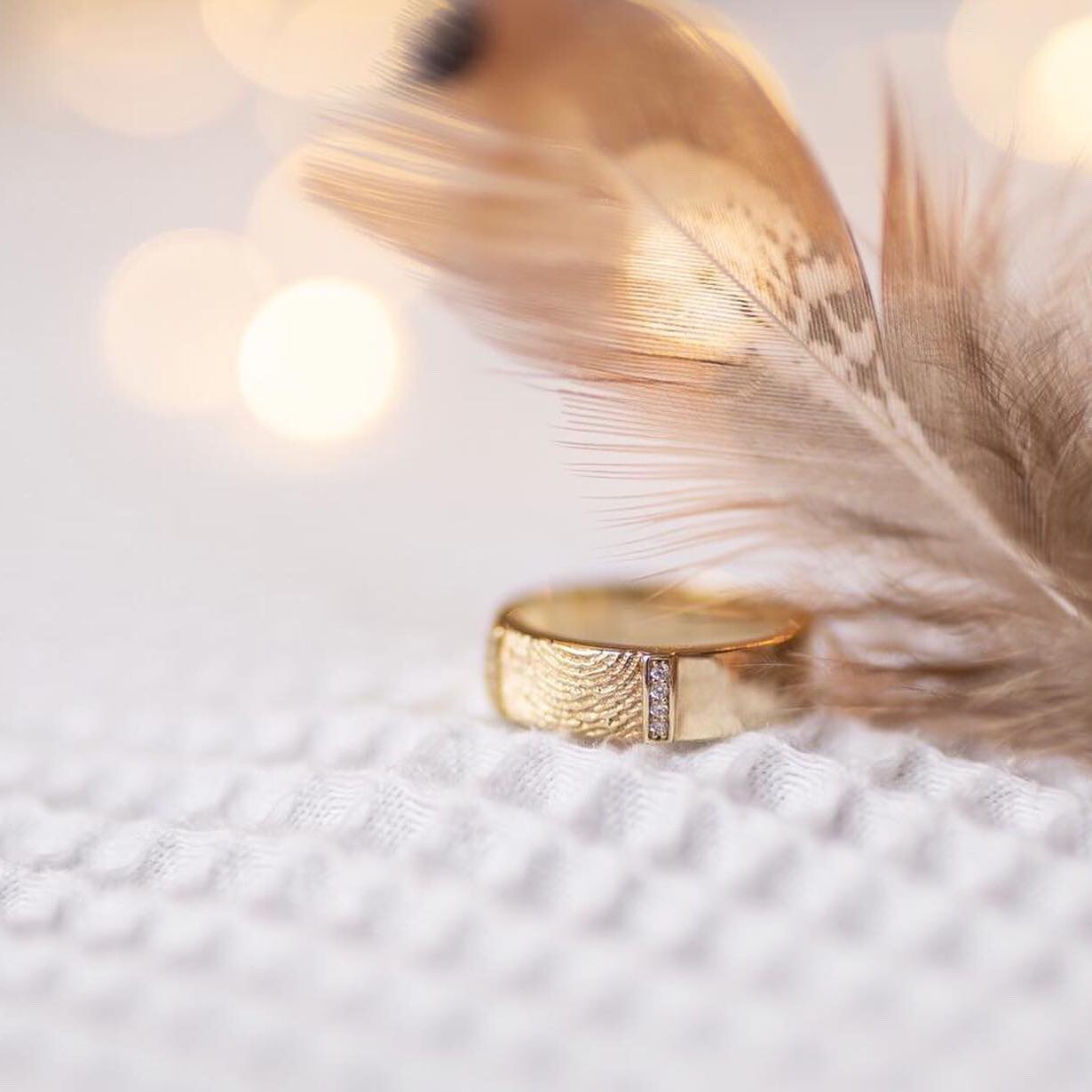 Gouden ring met kleine diamantjes naast een lichtbruine veer, tegen een wazige, zachtwitte achtergrond.