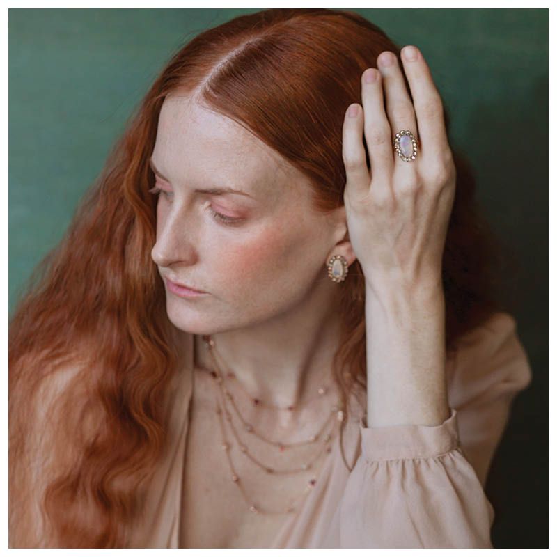 Vrouw met rood haar, sieraden dragend, kijkt met een peinzende blik naar beneden. Groene achtergrond.