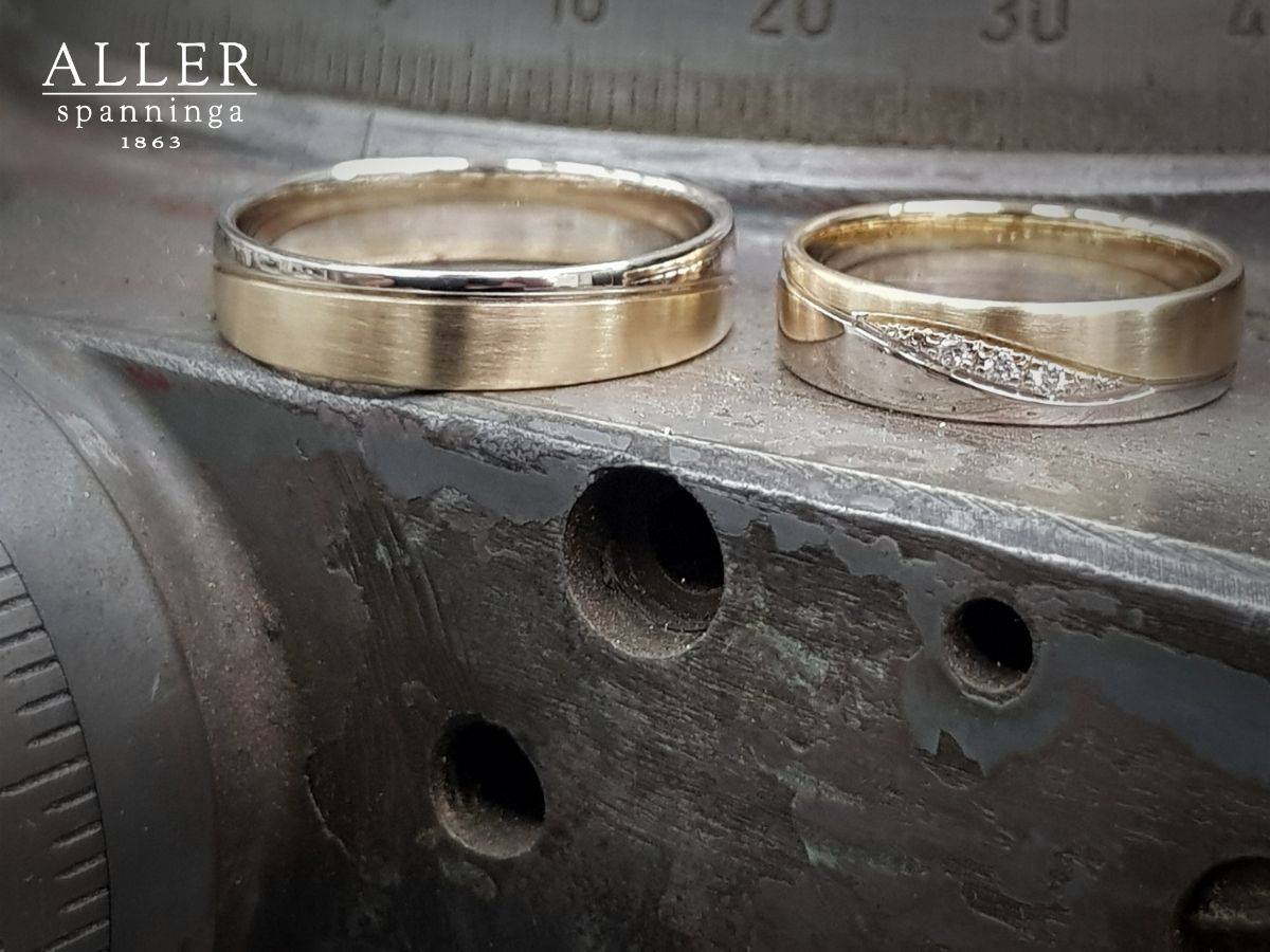 Twee gouden trouwringen op een metalen oppervlak, één met een diamantontwerp, de andere tweekleurig.