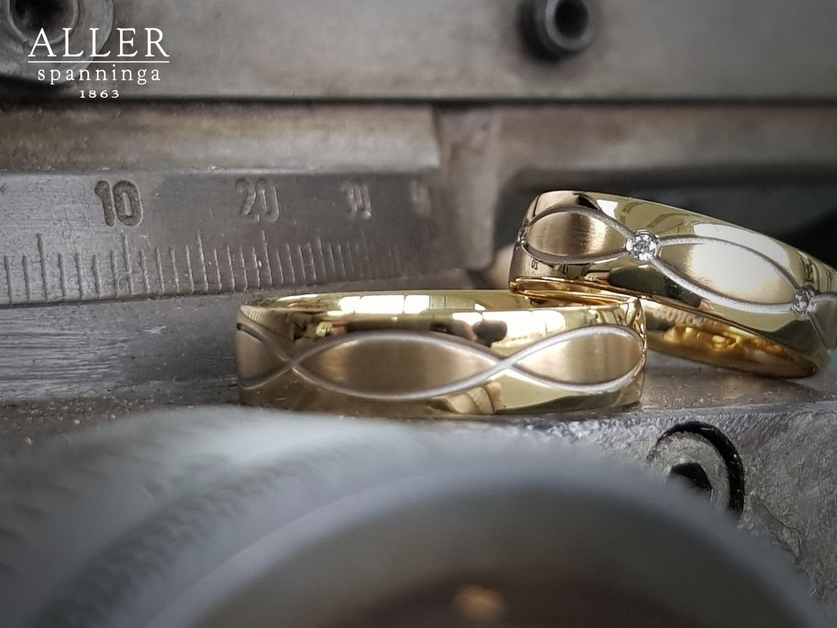 Twee gouden trouwringen met een gegraveerd ontwerp, rustend op een metalen oppervlak, tegen een vage industriële achtergrond.