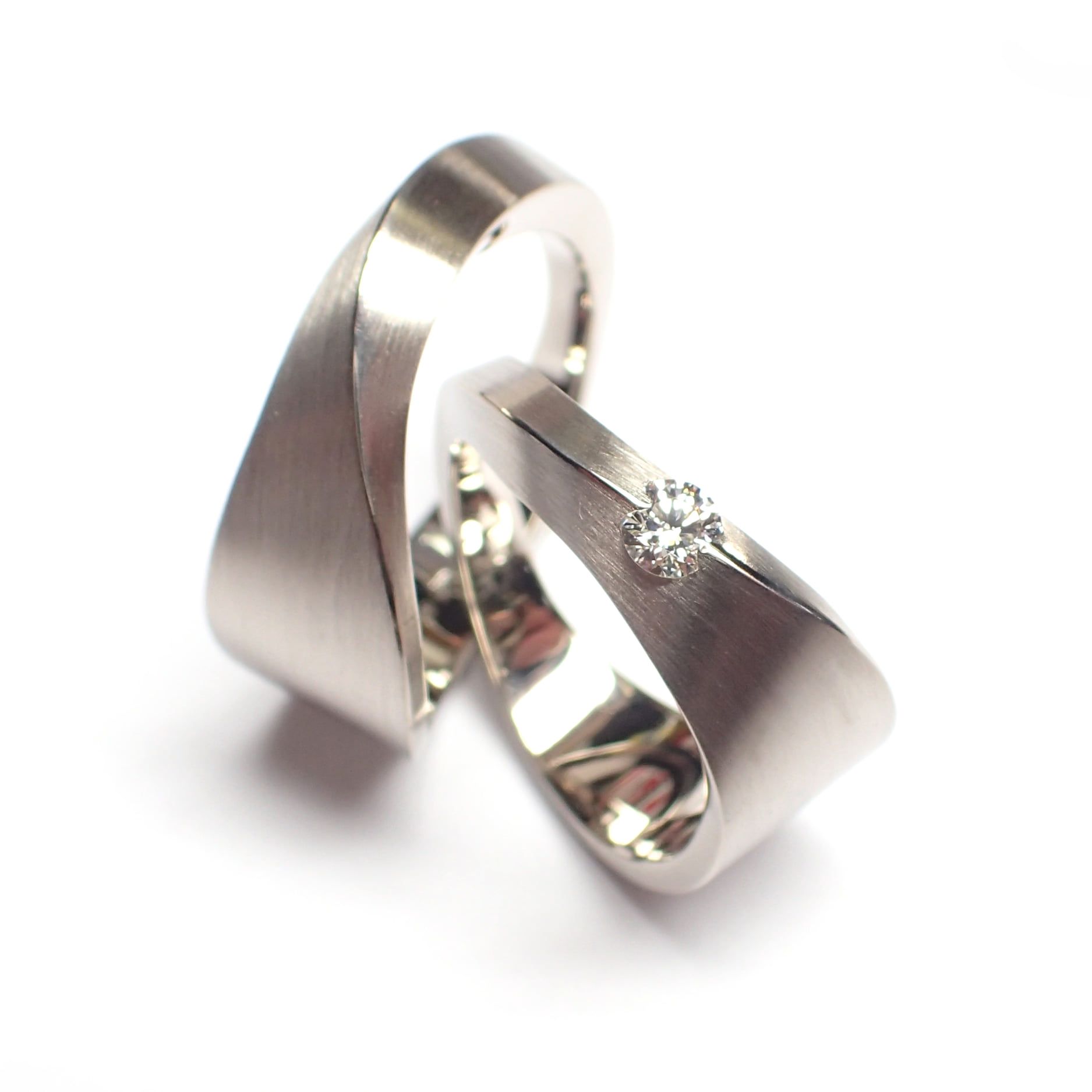 Twee zilveren trouwringen, waarvan één met een klein diamantje, op een witte achtergrond.