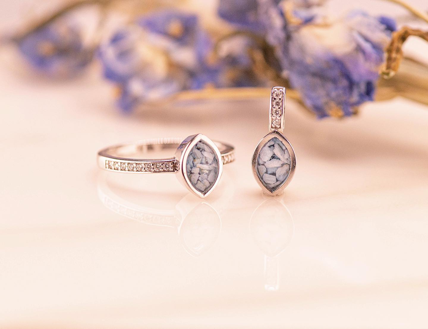 Ring en hanger bezet met een blauwgrijze markiesvormige steen, op een lichte ondergrond met gedroogde blauwe bloemen.