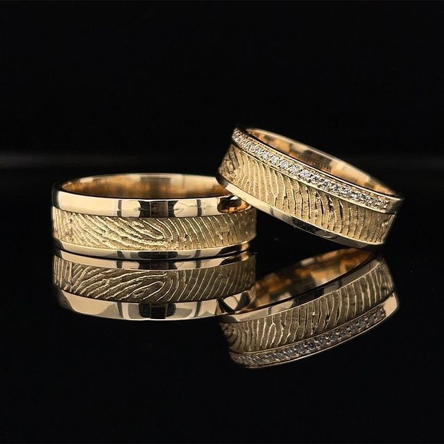 Twee gouden trouwringen met geëtste textuur en diamantaccenten, die reflecteren op een zwart oppervlak.