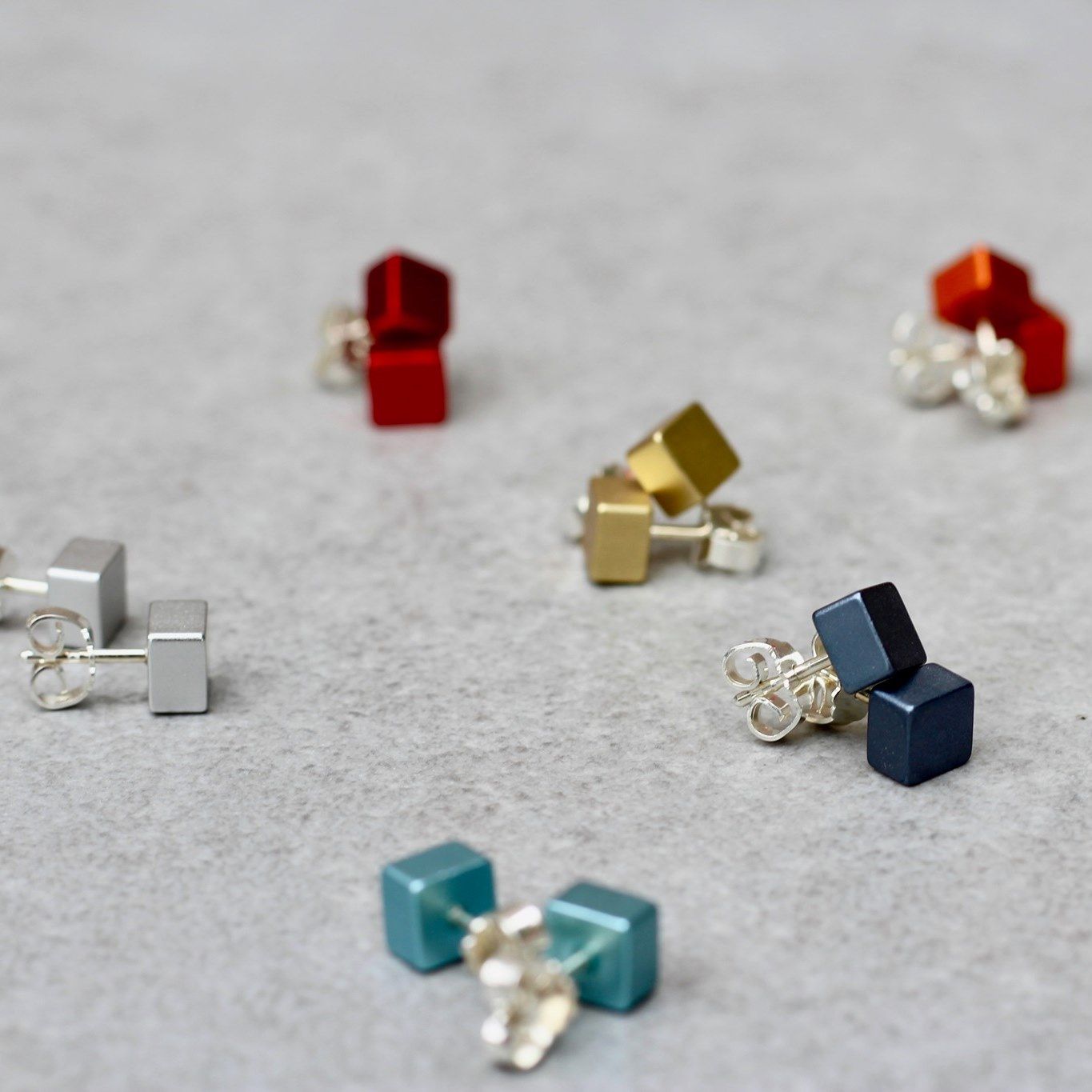 Kleurrijke, kubusvormige oorbellen verspreid op een grijs oppervlak; goud, rood, blauw, blauwgroen en zilver.