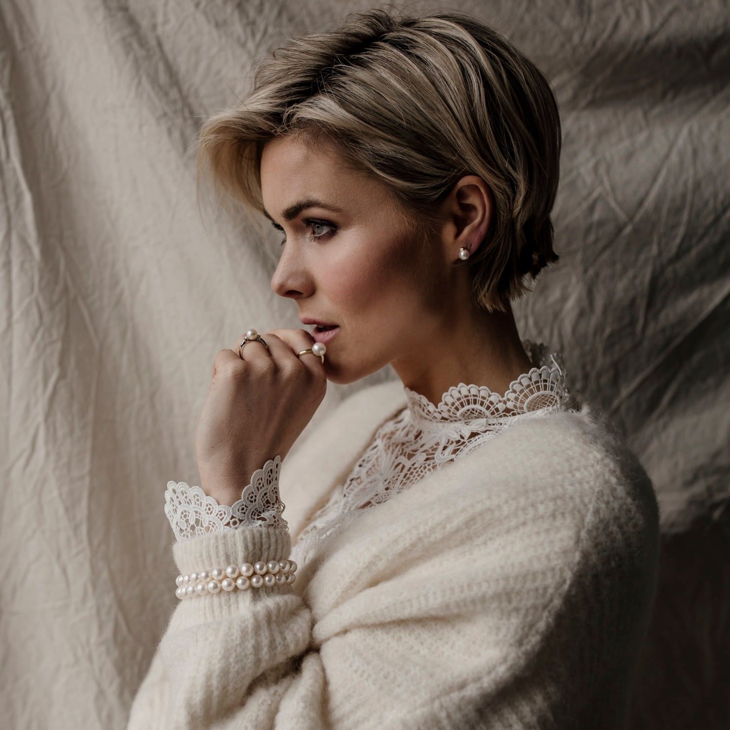 Vrouw met kort blond haar, gekleed in een crèmekleurige trui met kanten details, kijkt peinzend opzij.