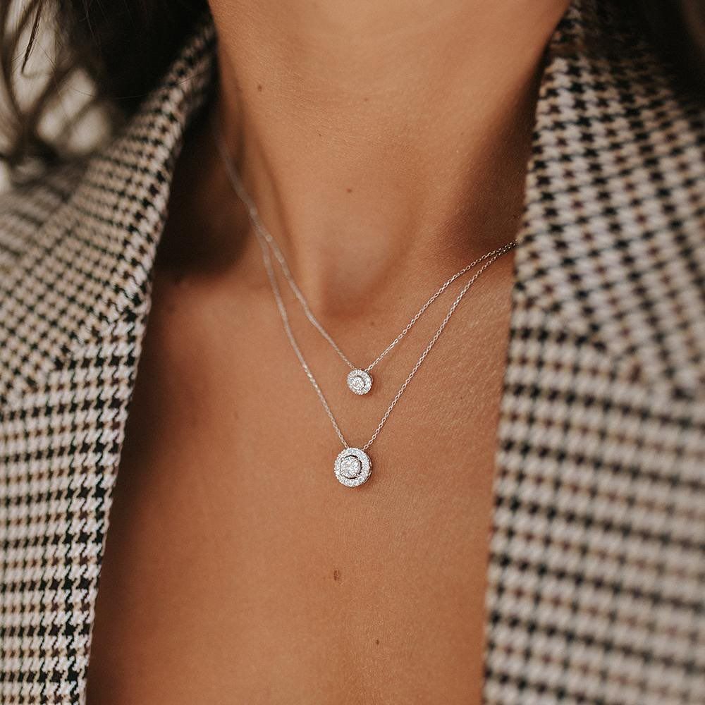 Twee kettingen met diamanten hangers om de nek van een vrouw, met een geruite blazer op de achtergrond.