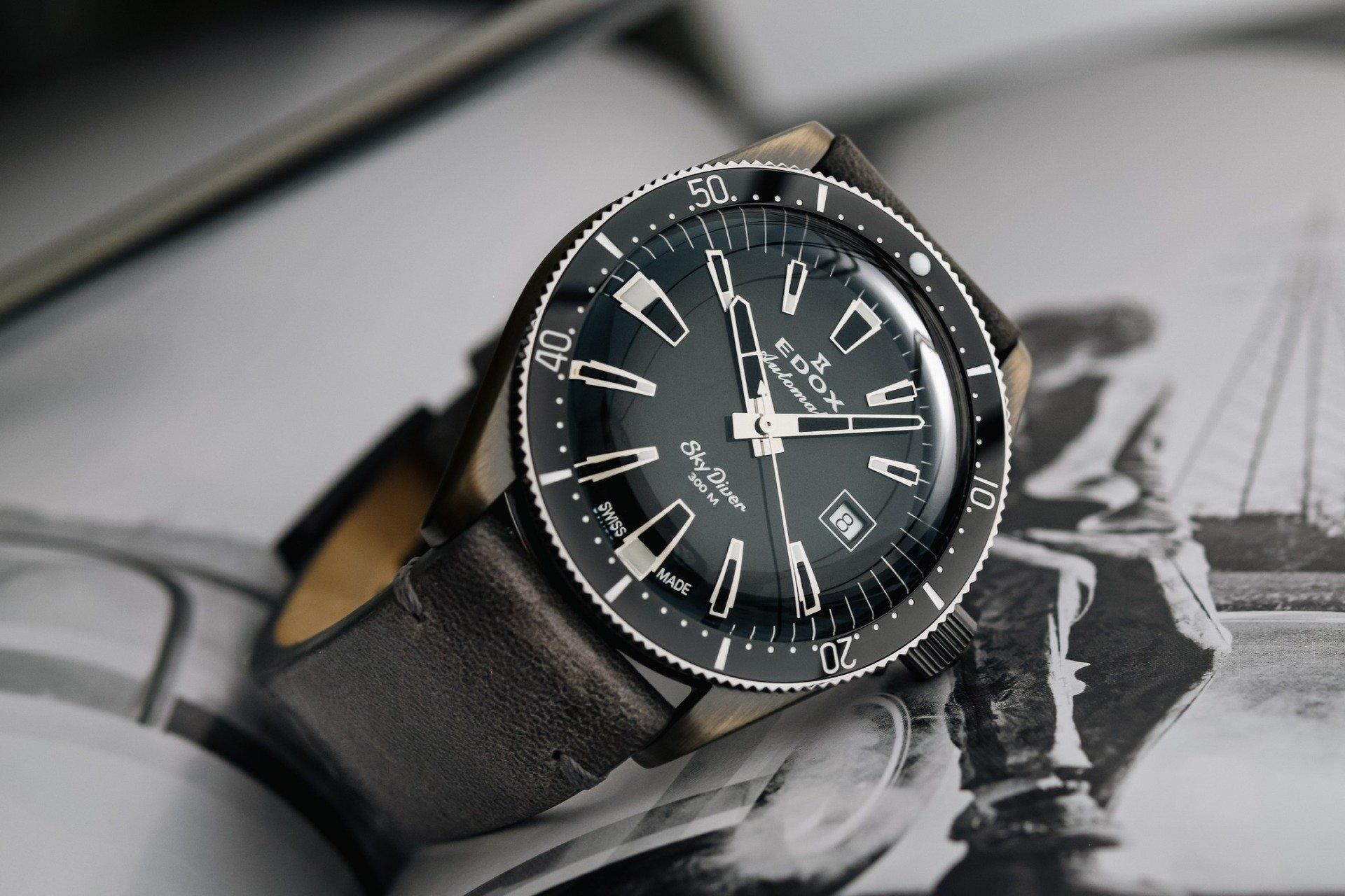 Close-up van een zwart horloge met een draaibare lunette en leren band op een zwart-witte achtergrond.