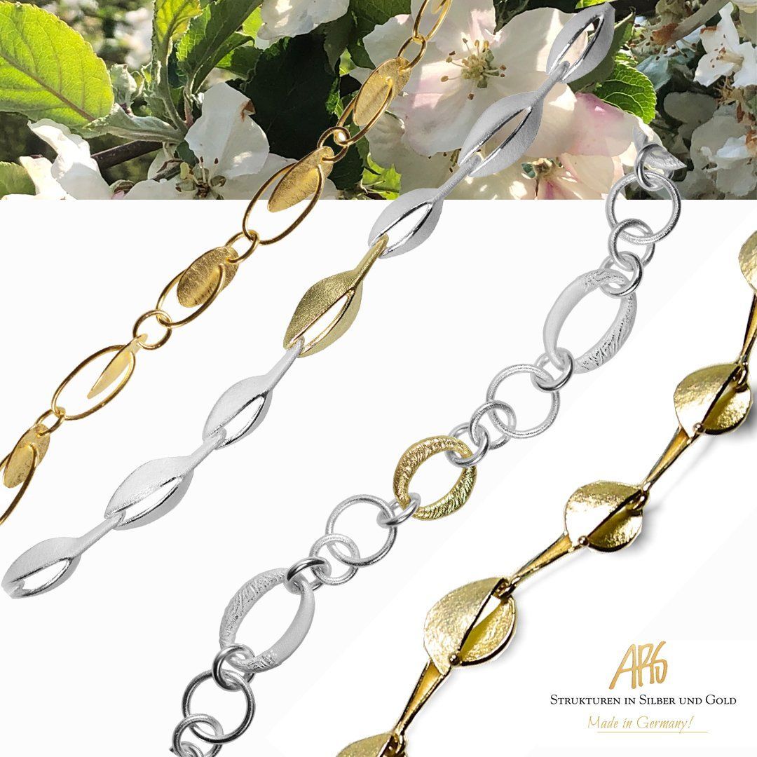 Close-up van een sieradenketting in goud en zilver, met bladeren en ovale schakels, tegen een witte bloemenachtergrond.