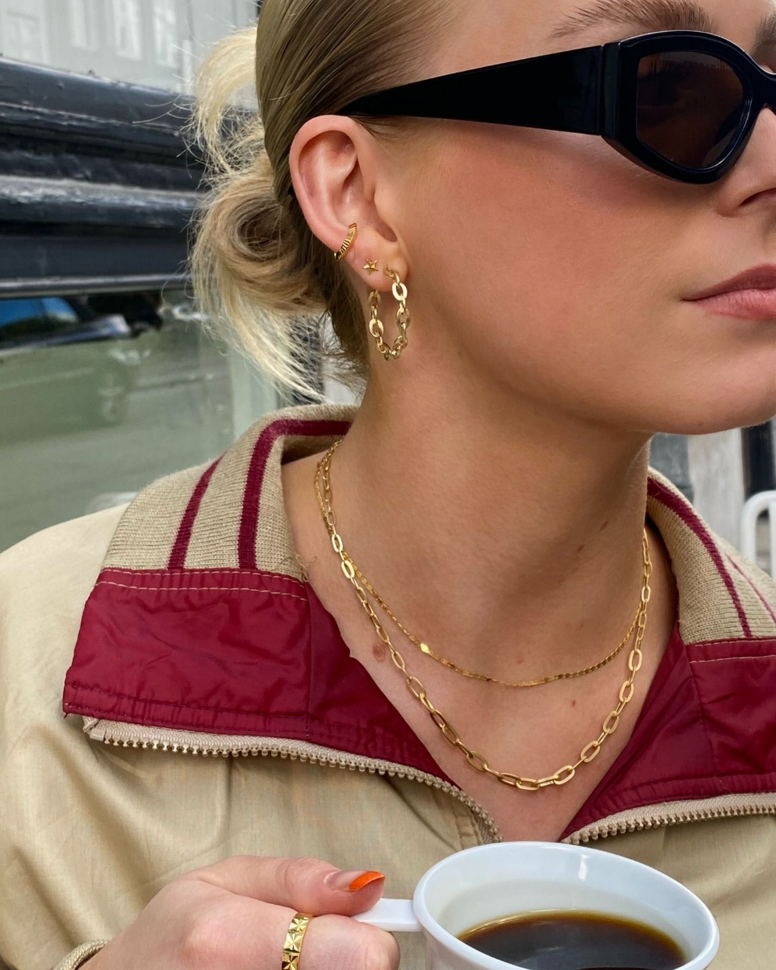 Vrouw met zonnebril, gouden sieraden en een beige jas, met een mok koffie in haar hand.