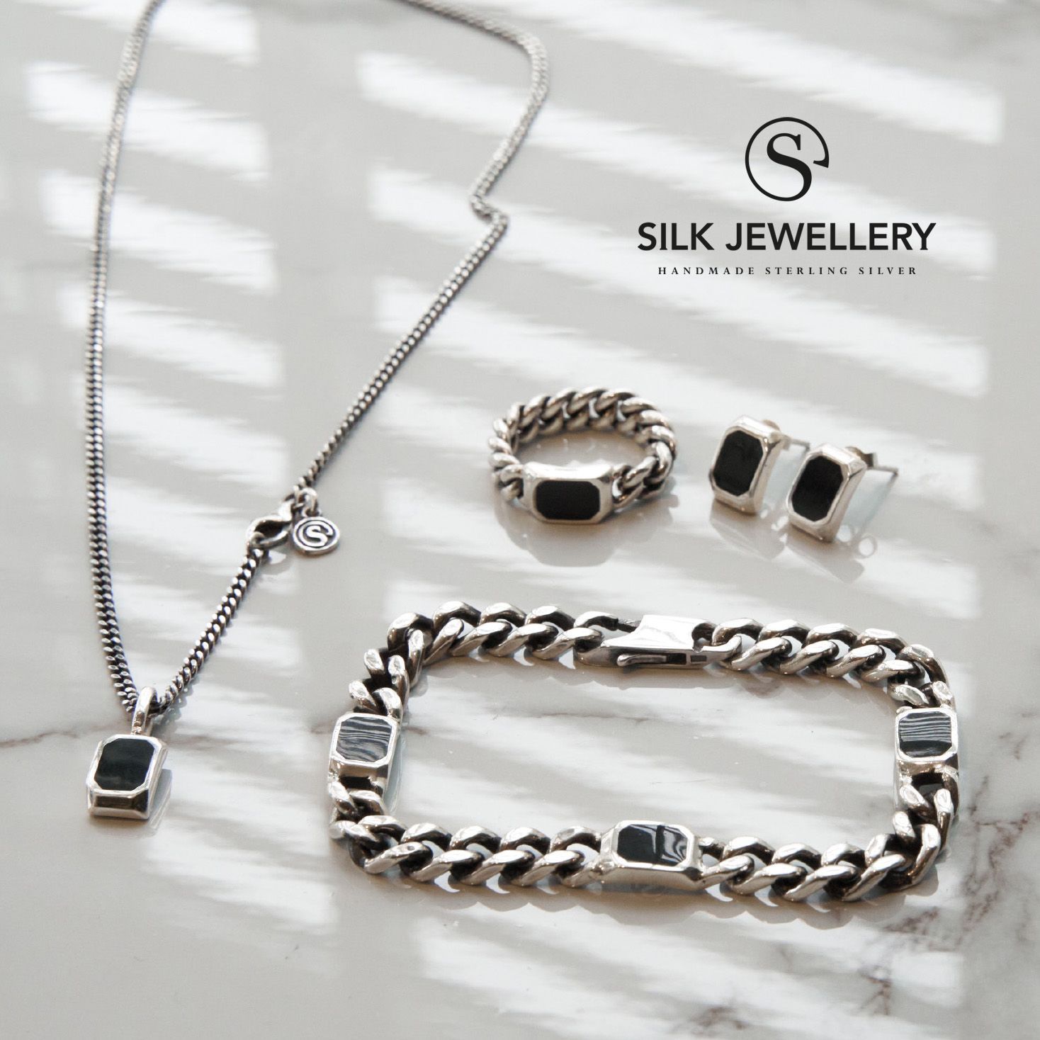 Zilveren sieraden op een marmeren ondergrond: ketting, ring, armband en oorbellen met zwarte accenten.
