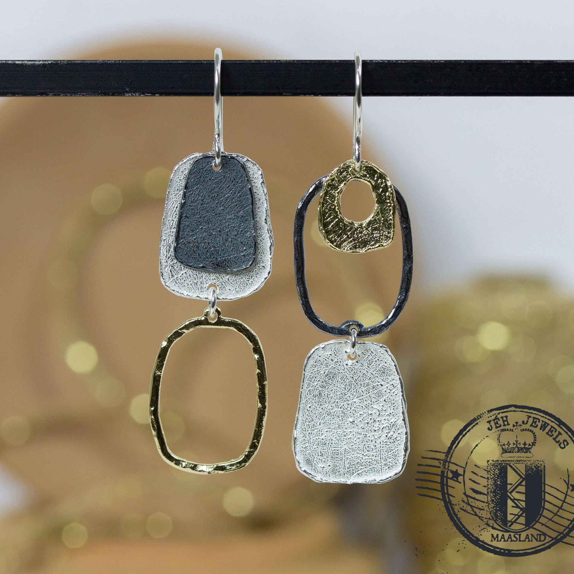 Abstracte oorbellen van zilver en goud met een getextureerd oppervlak, hangend aan een zwarte staaf.