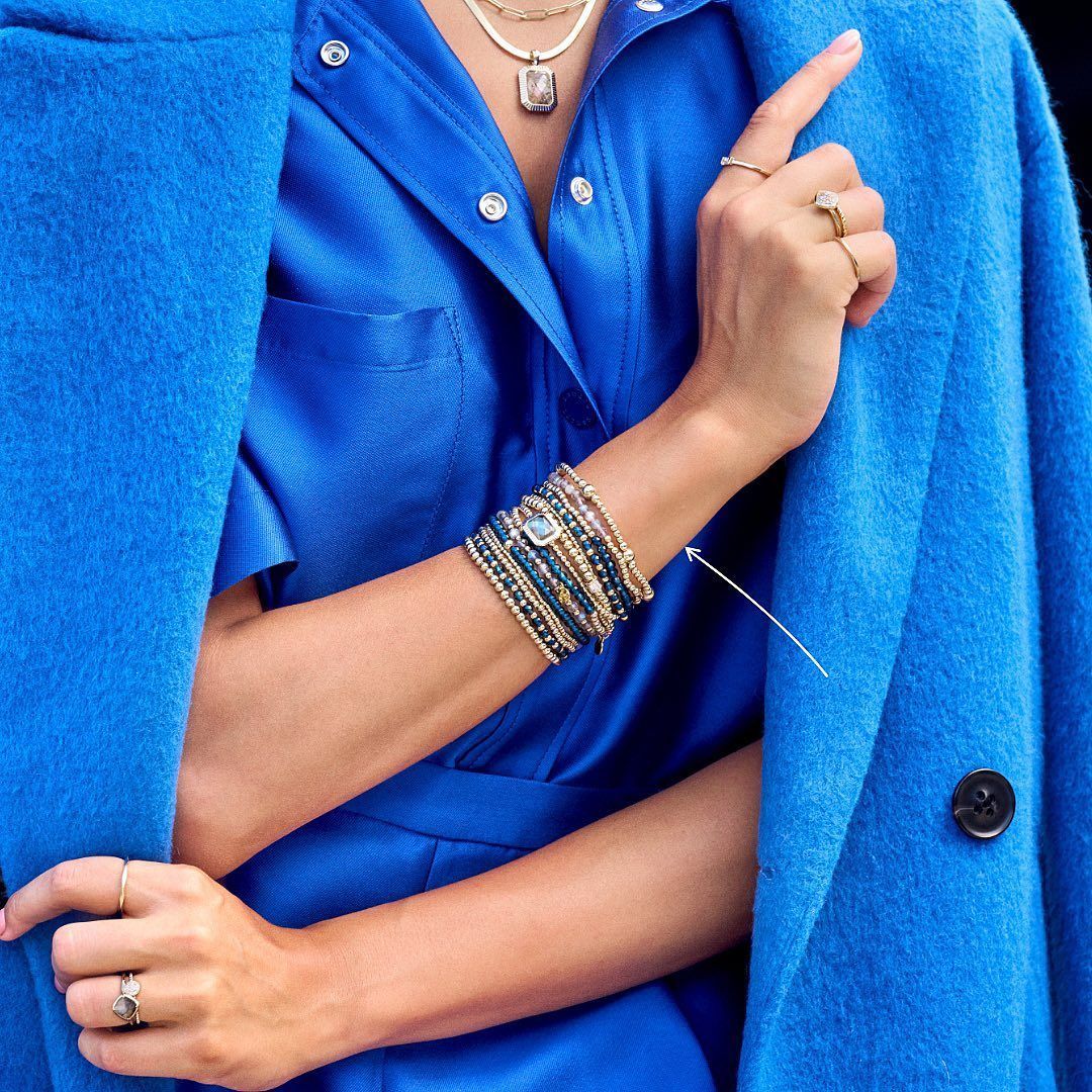 Vrouw in blauwe outfit, met ringen en armbanden, met een blauwe jas over haar schouders.